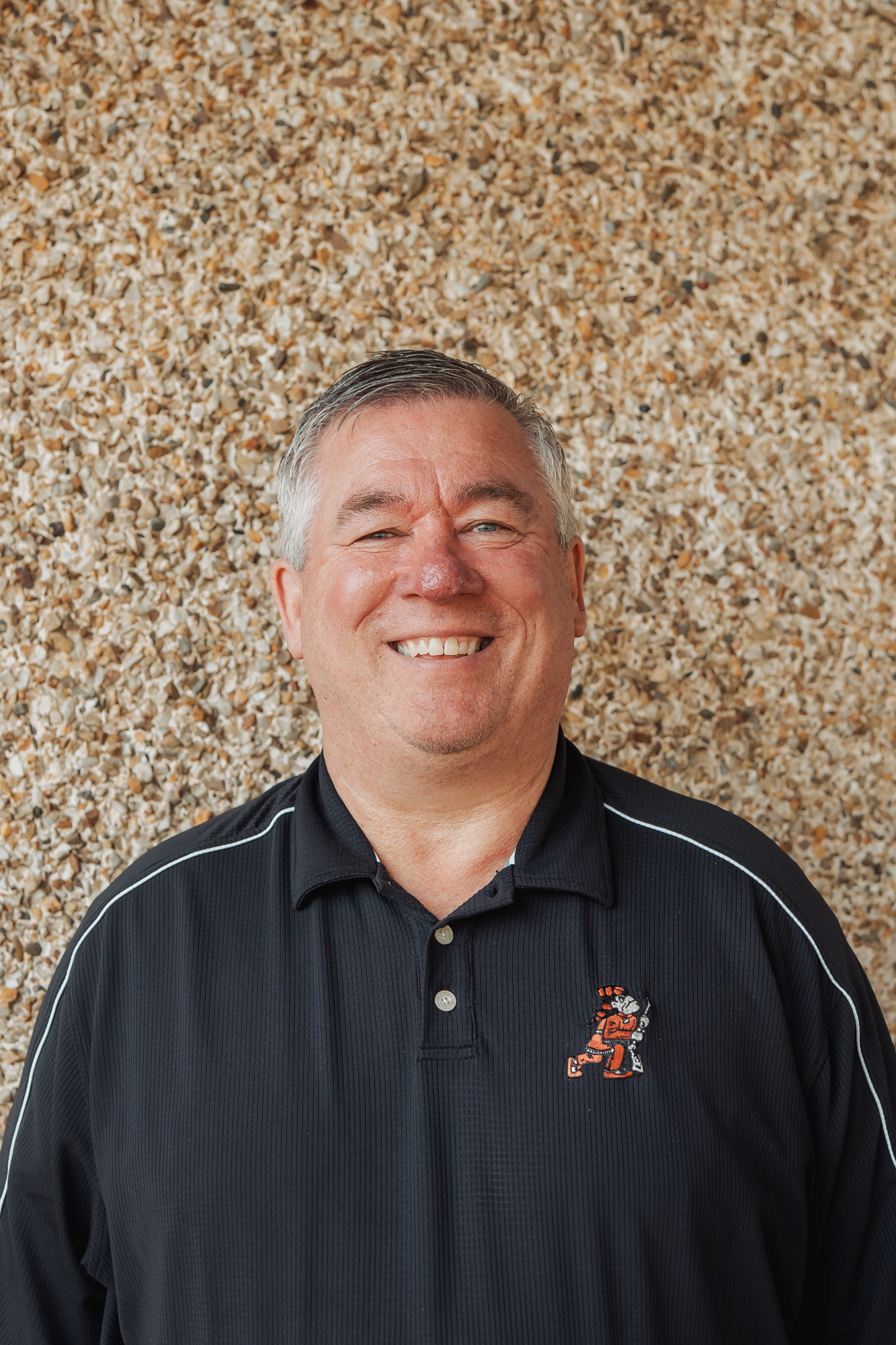 A smiling man in a black polo shirt stands in front of a textured wall.