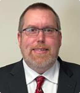 A man with glasses and a beard smiles, wearing a suit and tie.