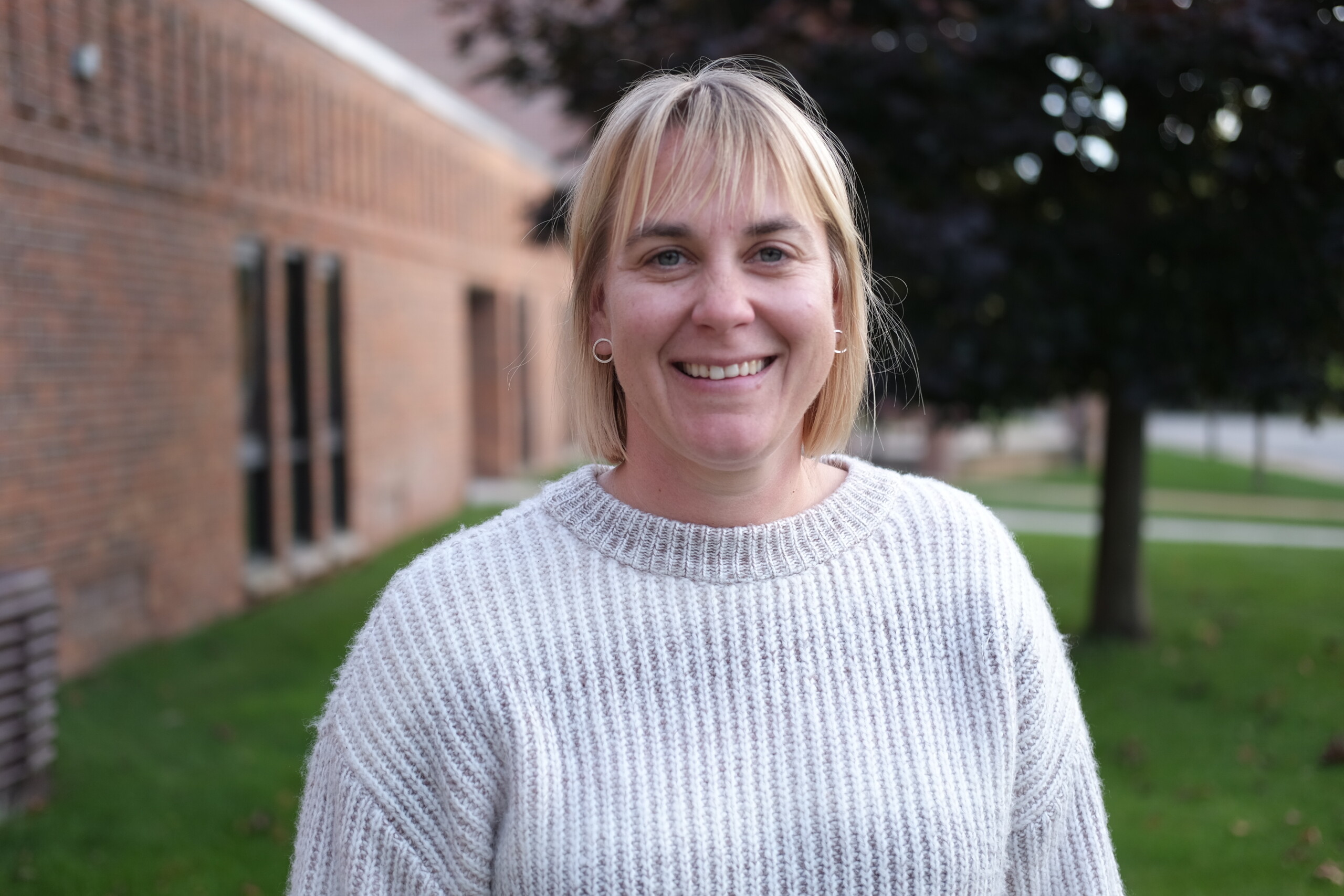 A woman with blonde hair and a white sweater smiles outside a brick building.
