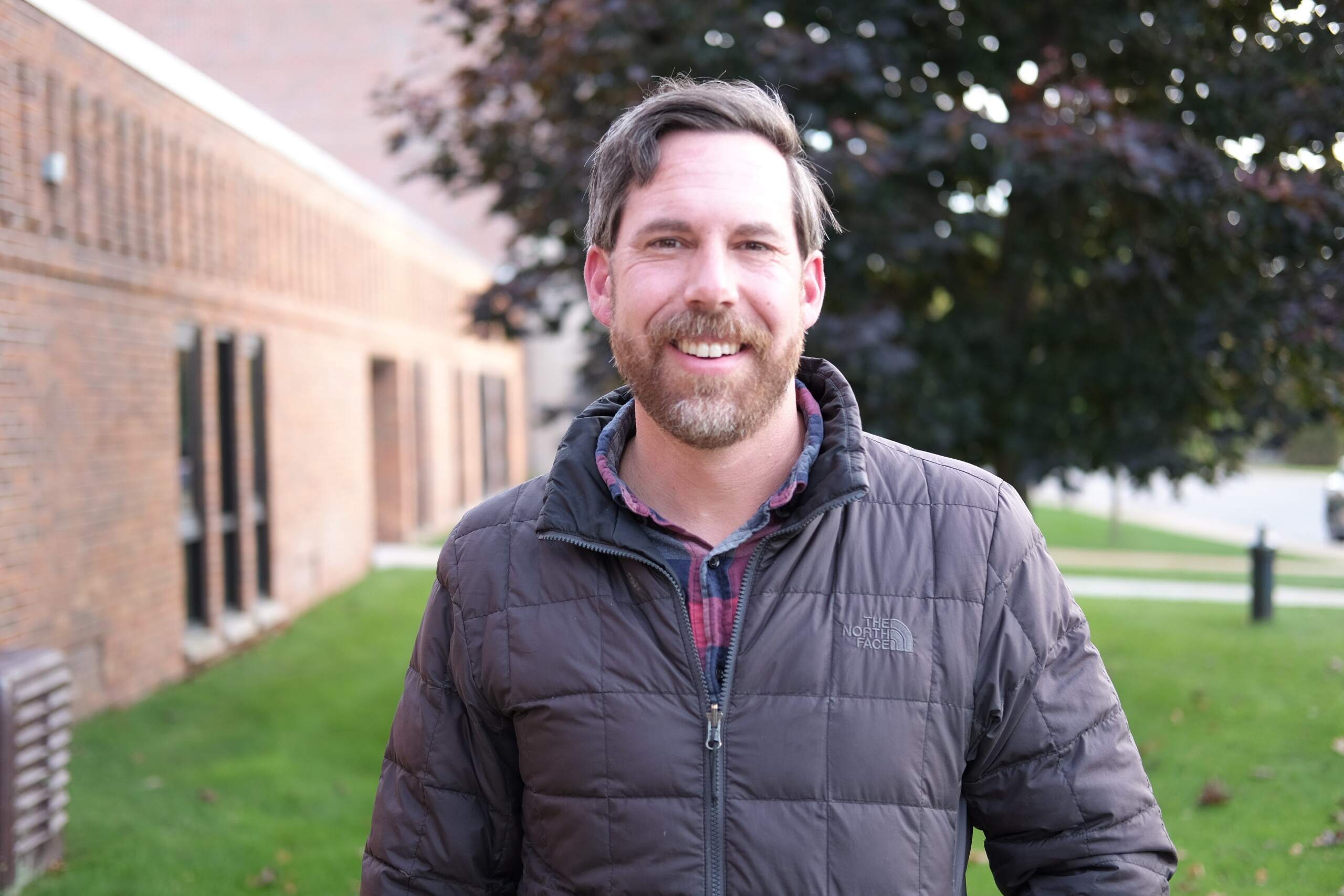 A smiling man with a beard stands outdoors near a brick building and green grass.