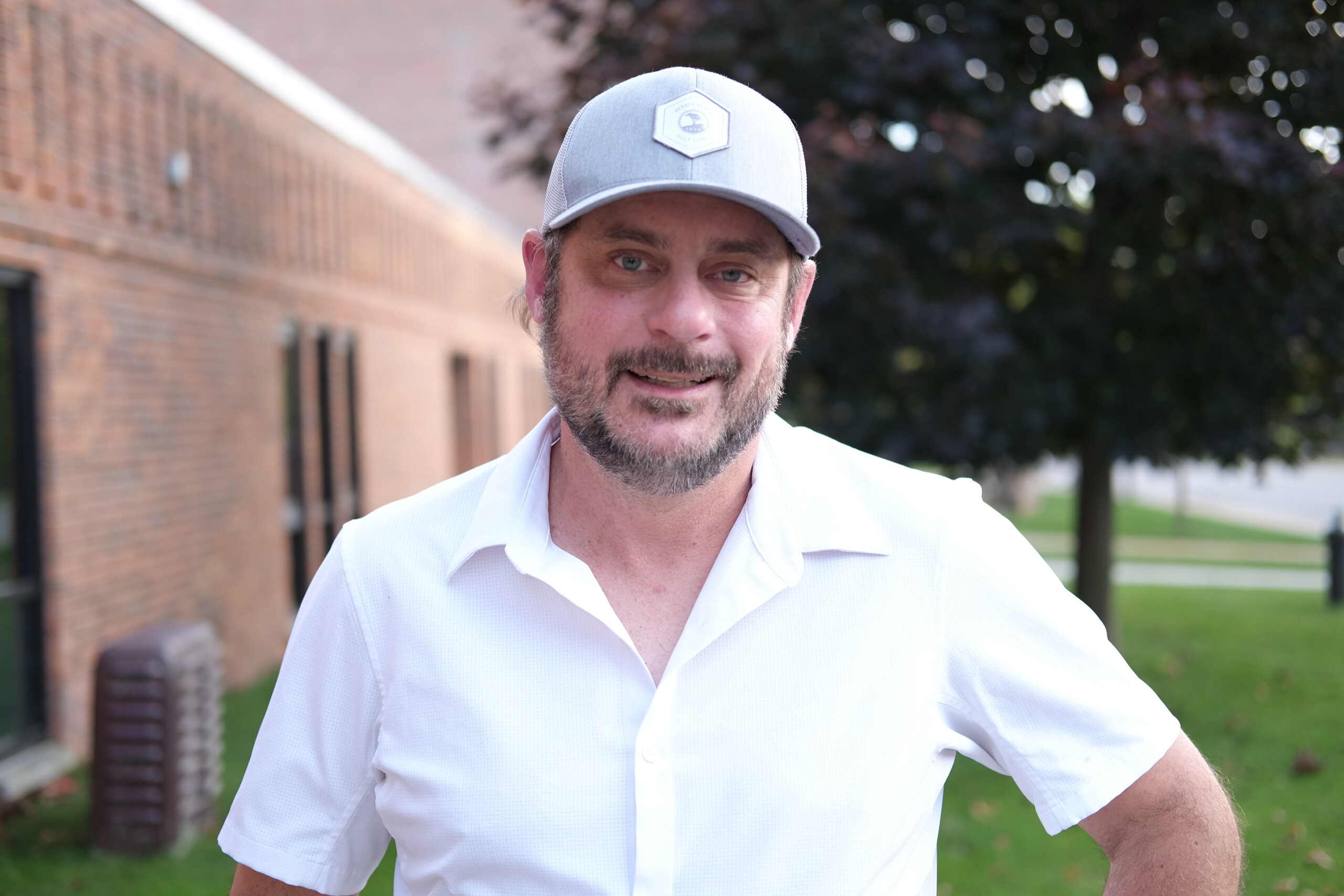 A smiling man wearing a white shirt and a grey baseball cap stands outdoors near a brick building.