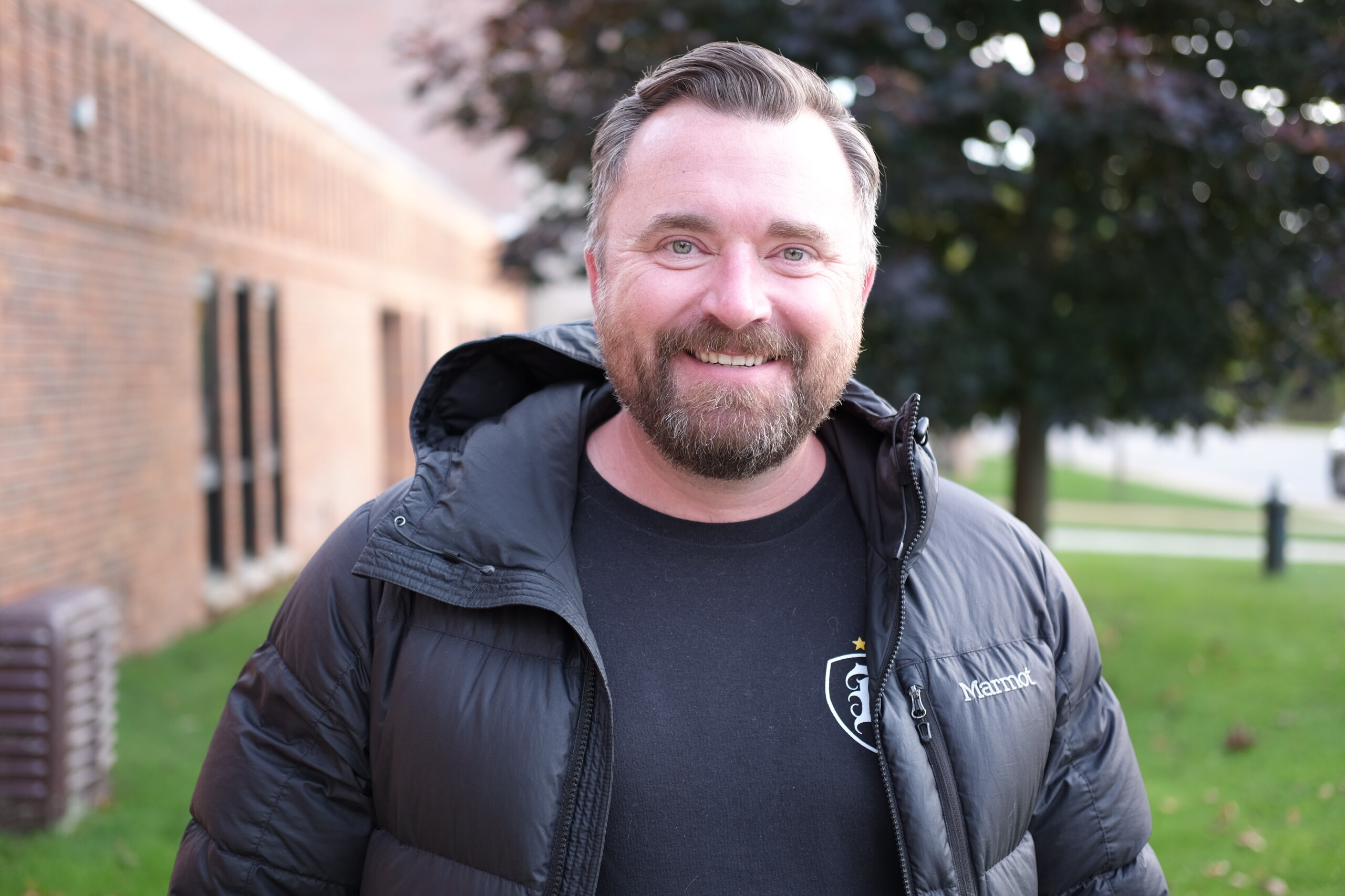A smiling man with a beard stands outside, wearing a black puffer jacket and t-shirt.