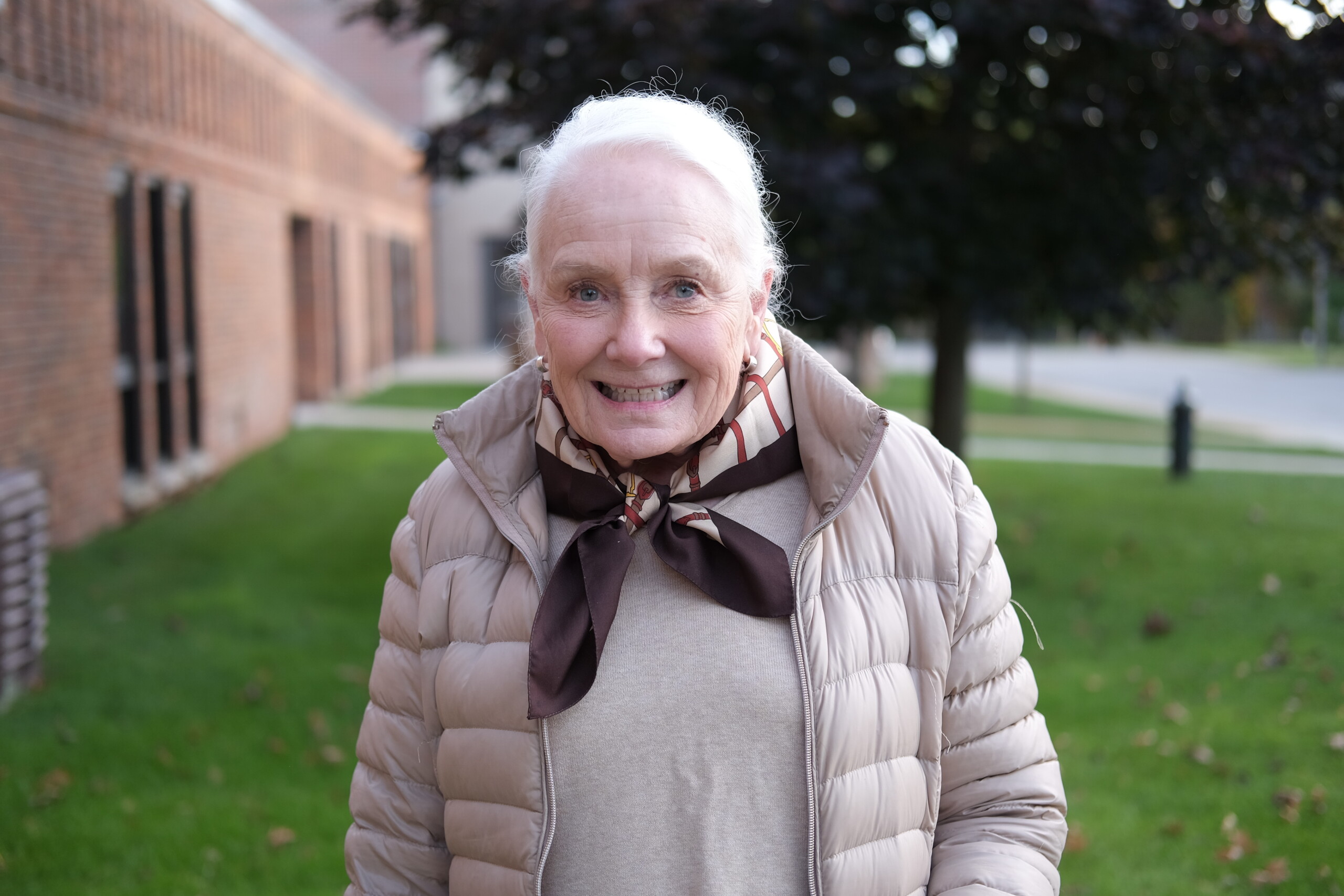 A smiling older woman with white hair stands outside, wearing a beige puffer jacket and a brown patterned scarf.