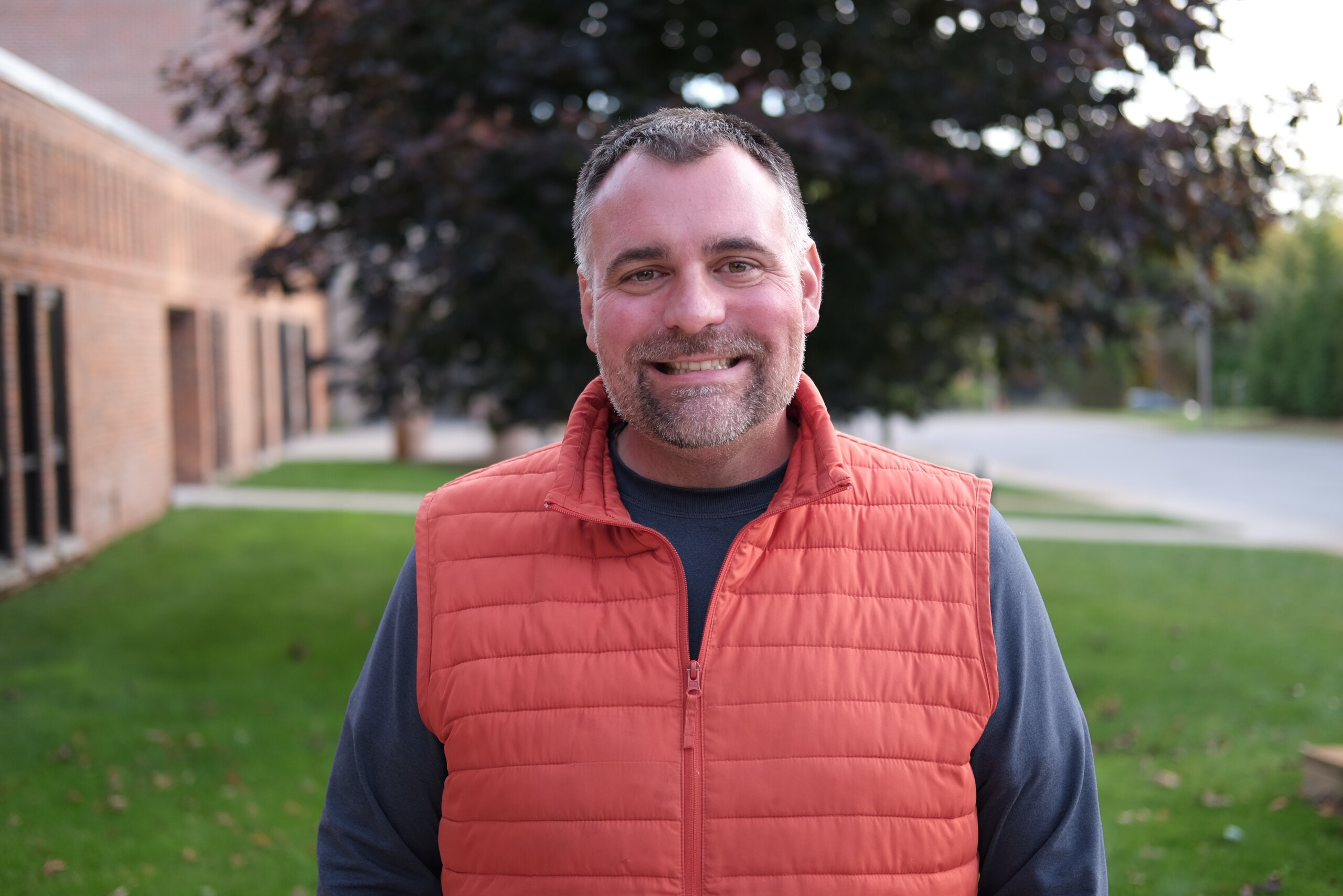 A smiling man stands outdoors wearing an orange puffer vest over a dark long-sleeve shirt.