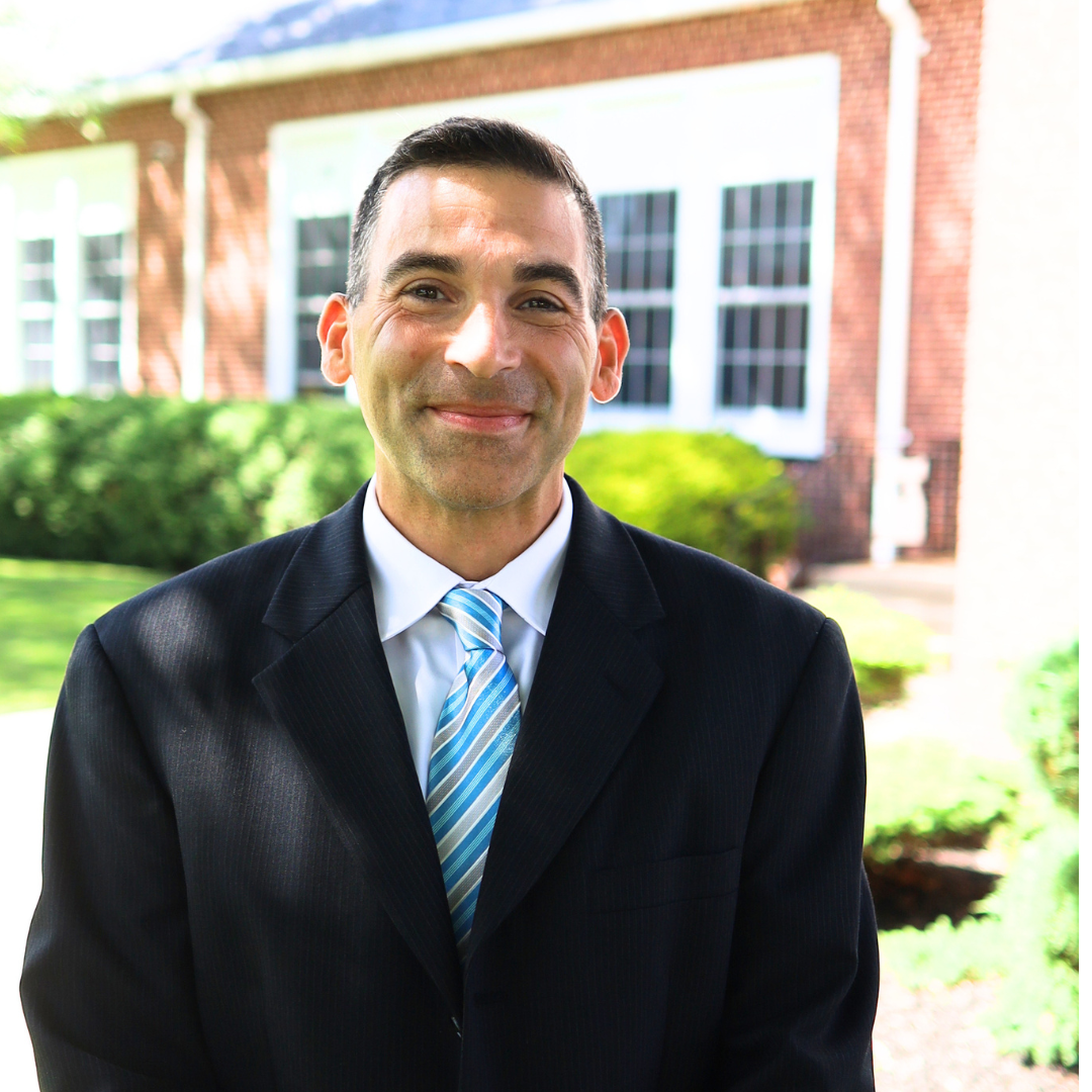 A man in a dark suit and striped tie smiles at the camera outdoors.