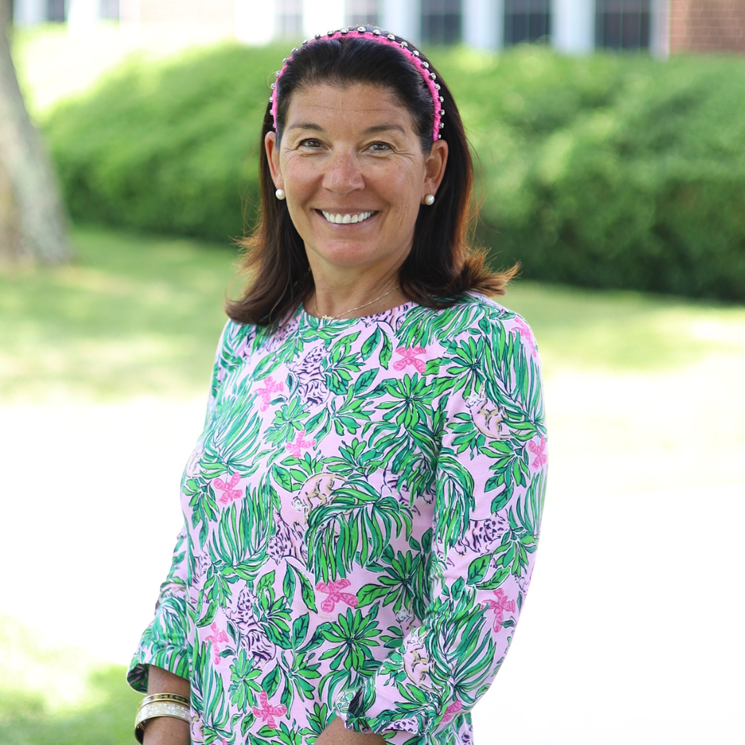 A smiling woman with dark hair and a pink headband wears a colorful tropical print dress.