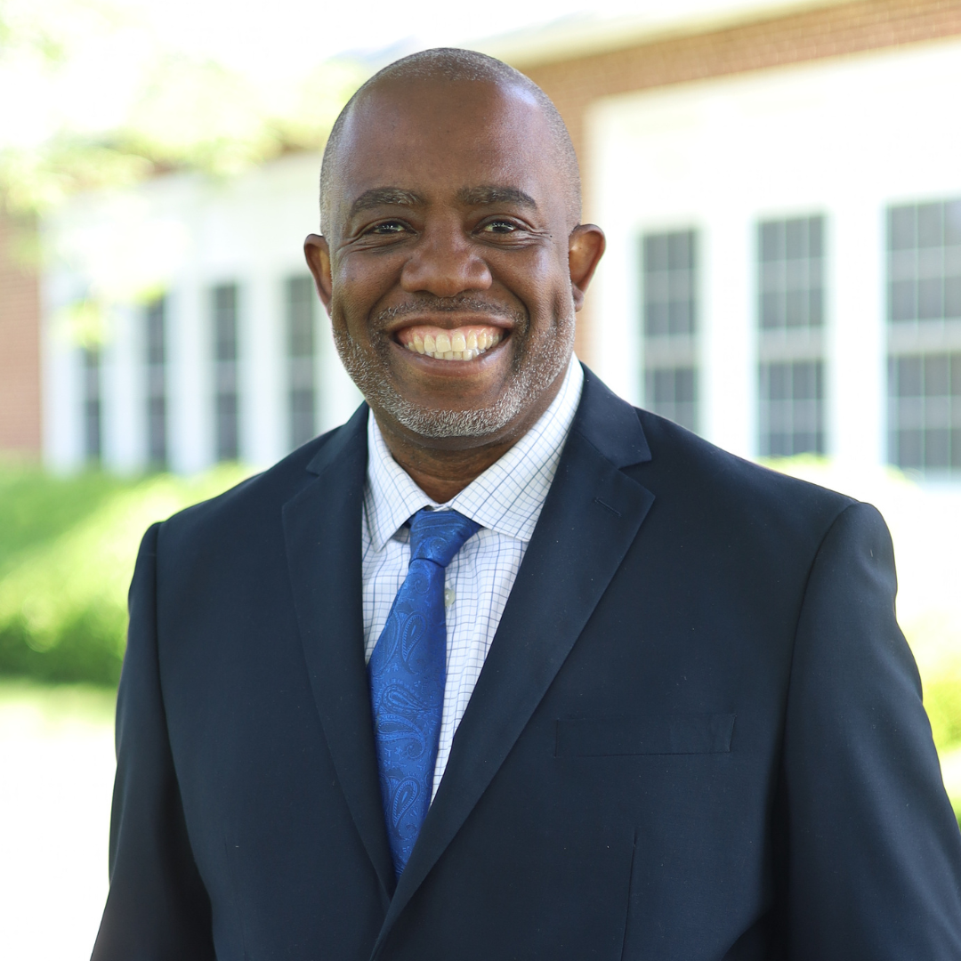 A smiling man in a dark suit and blue tie stands outdoors.