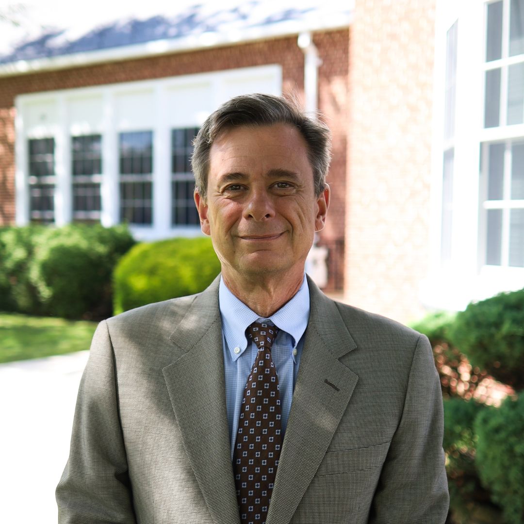 A man in a suit and tie smiles at the camera, standing in front of a brick building.
