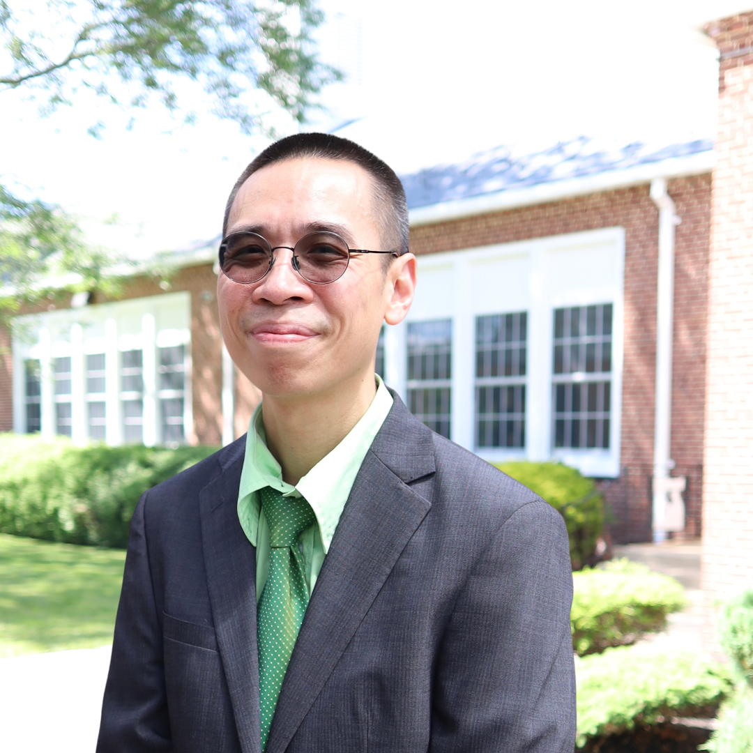 A man in a suit and tie stands outdoors in front of a brick building.