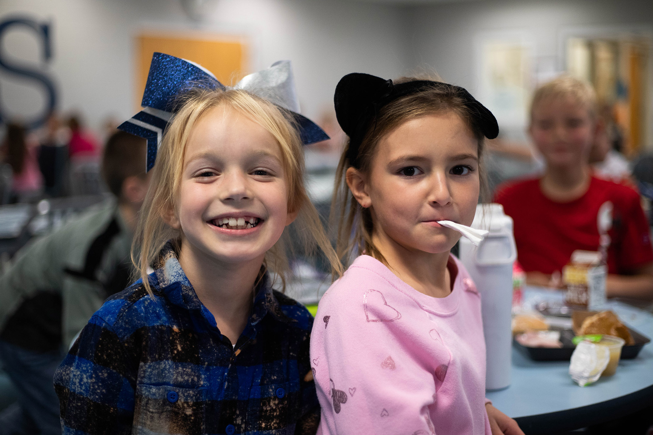 Two young girls smile at the camera in a school cafeteria.