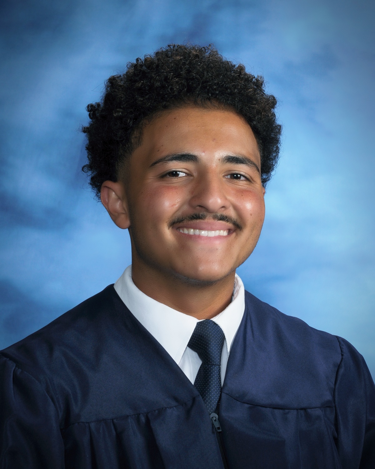 A young man in a graduation gown smiles warmly against a blue backdrop.