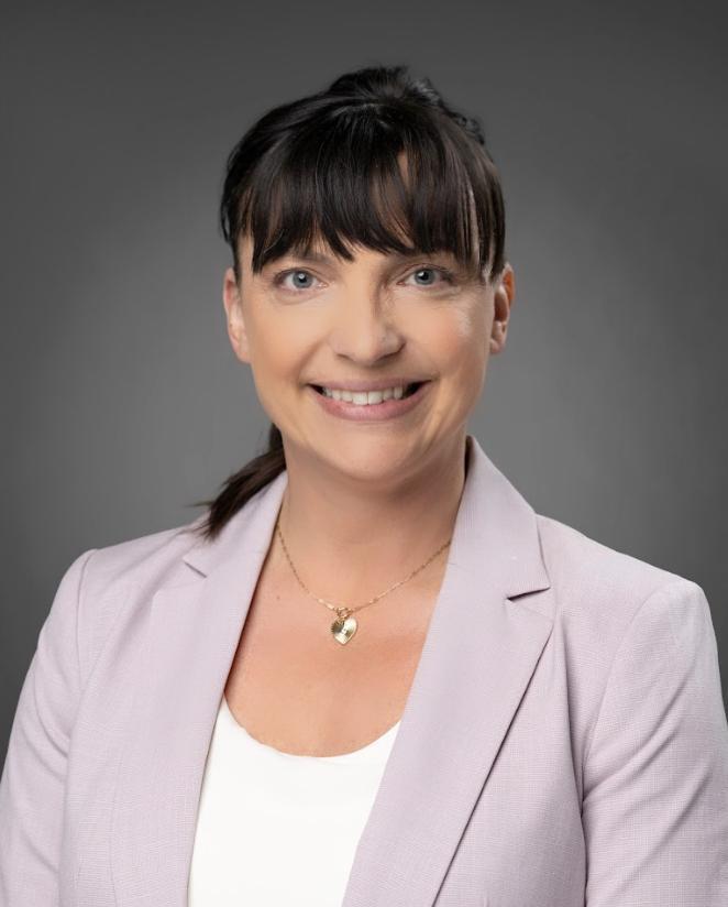A professional headshot of a smiling woman with dark hair pulled back, wearing a light purple blazer.