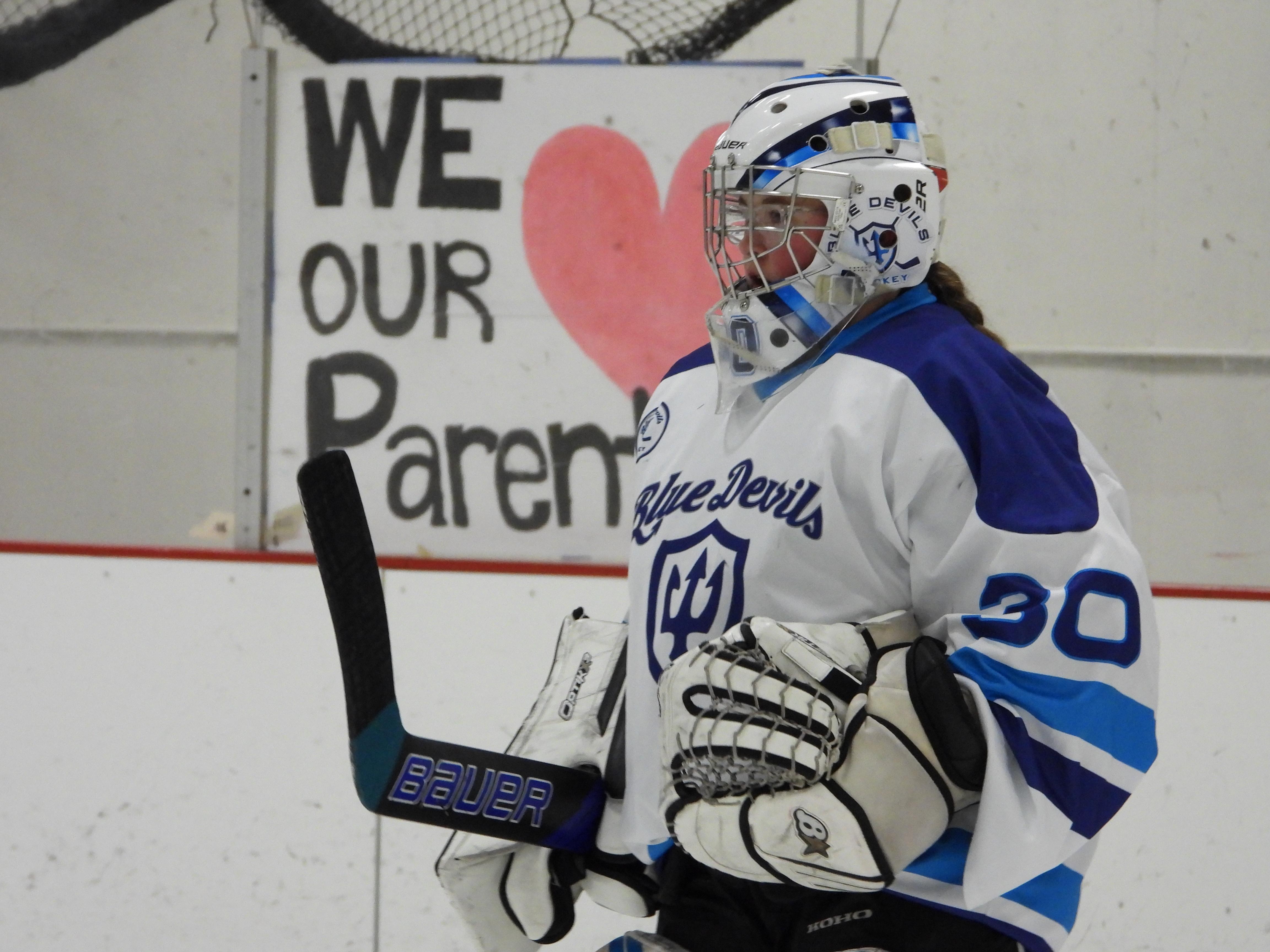 A hockey goalie in a blue and white uniform stands on the ice, holding a stick.