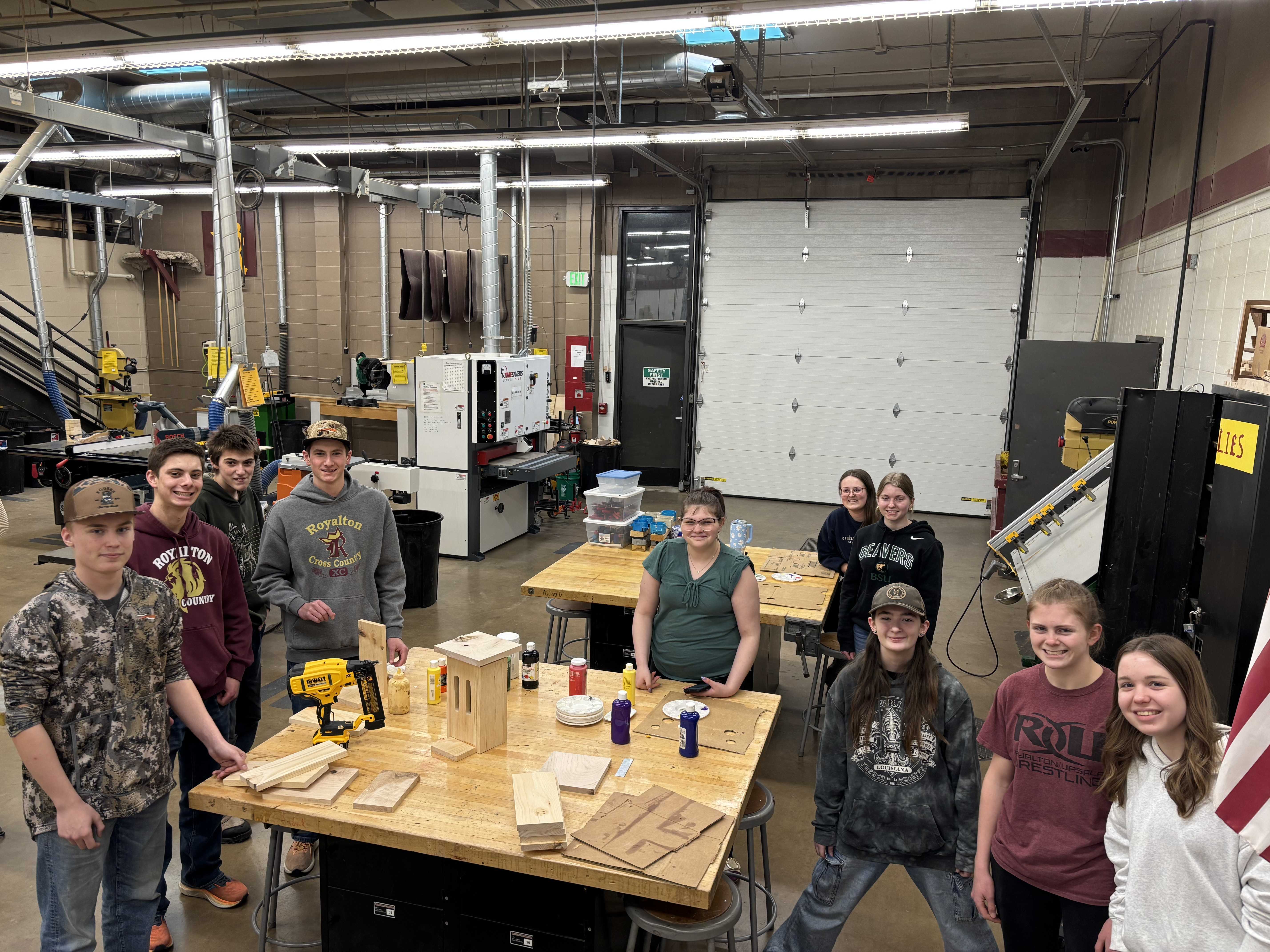 A group of people stand in a workshop, posing near wooden projects.