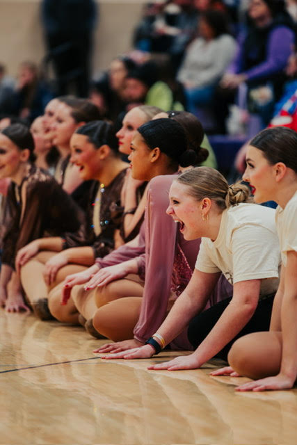 A group of young dancers watch intently from the sidelines of a competition.