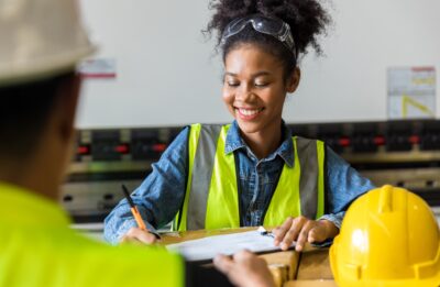 A smiling young woman in a hard hat and safety vest reviews a document.
