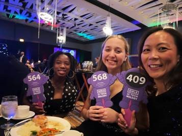Three smiling women hold up auction paddles with numbers 564, 568, and 409 at an event.