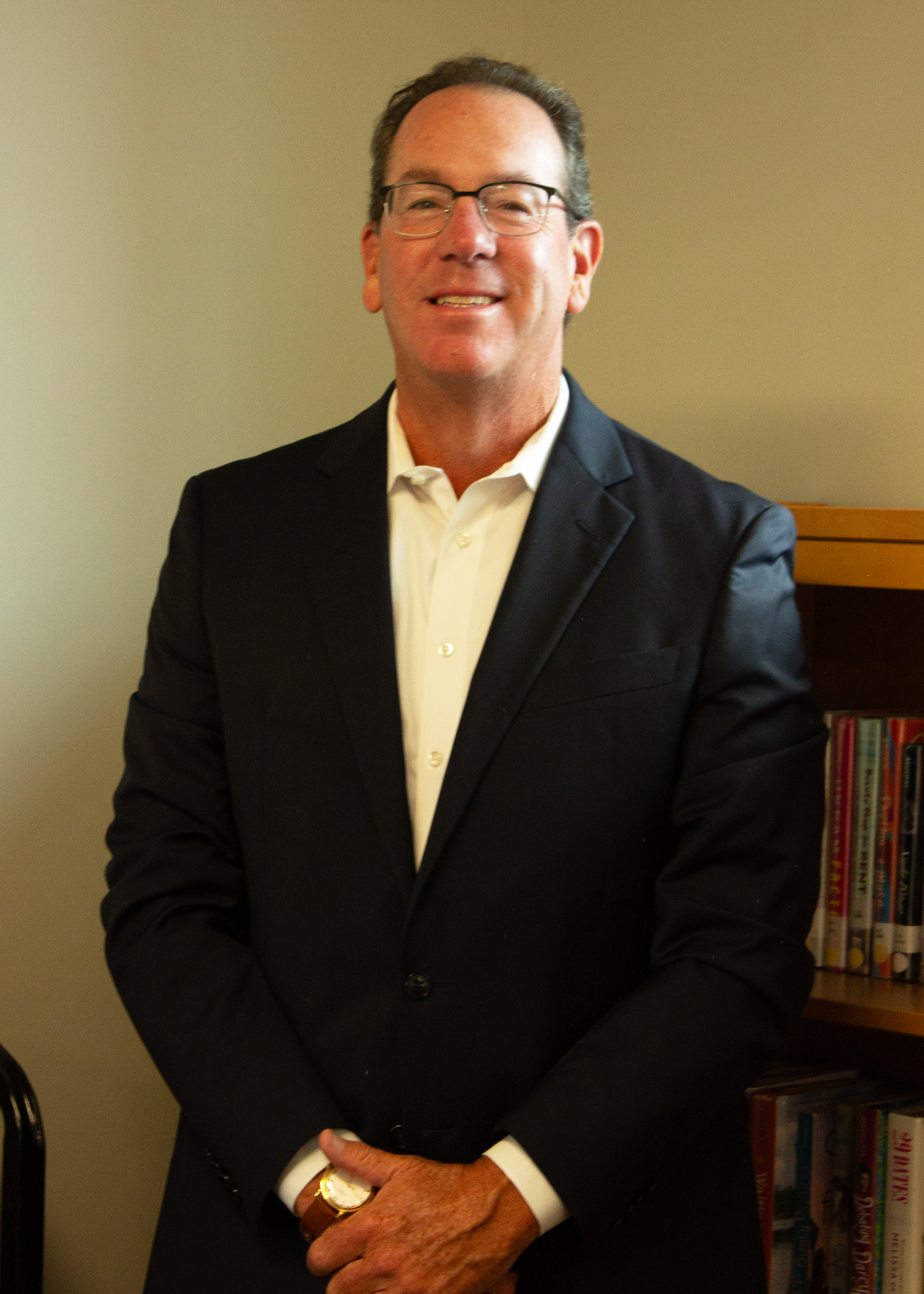 A man in a dark suit and glasses smiles, standing in front of a bookshelf.