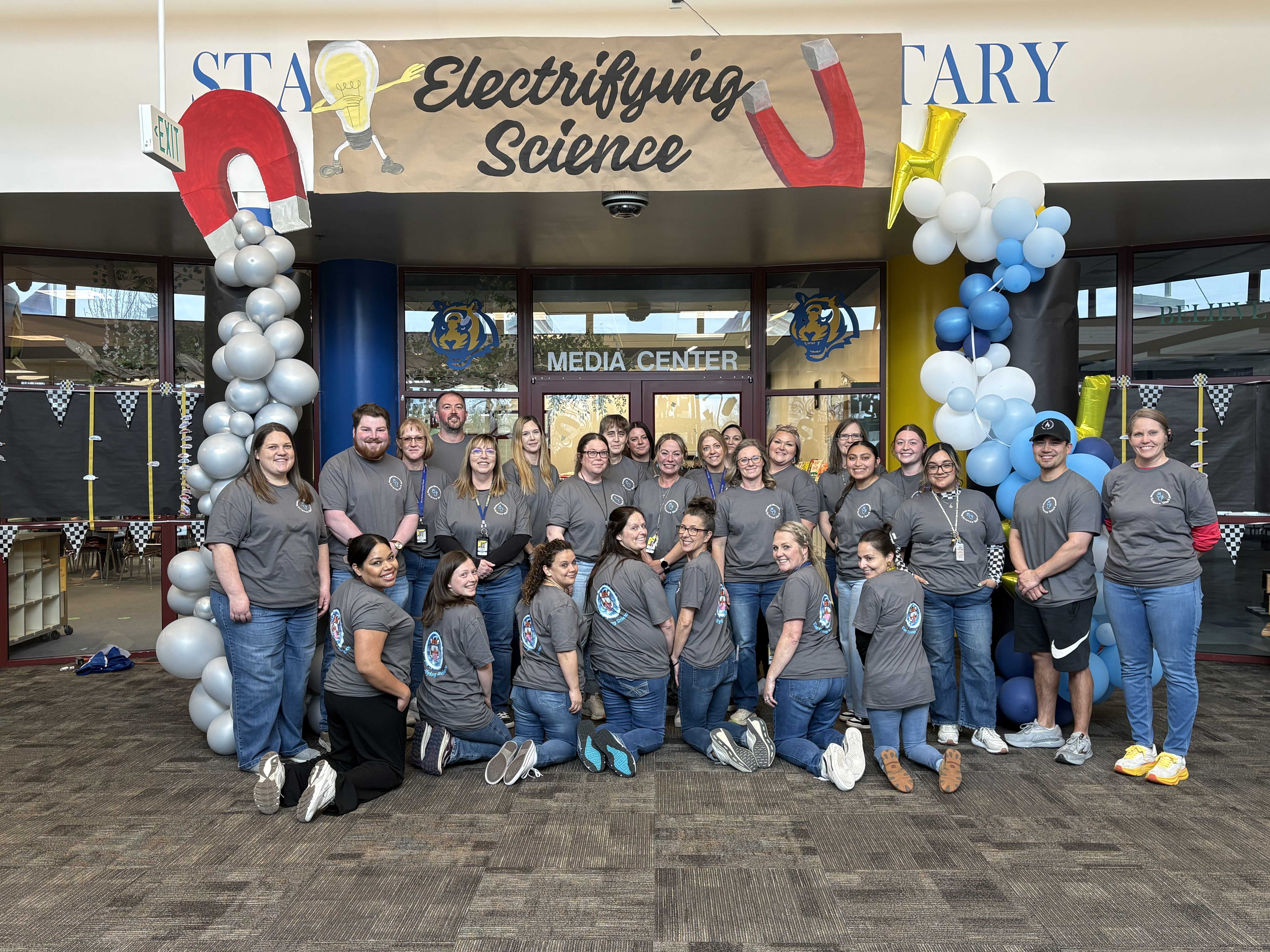 A group of people in matching t-shirts pose for a photo under a banner that reads 'Electrifying Science'.