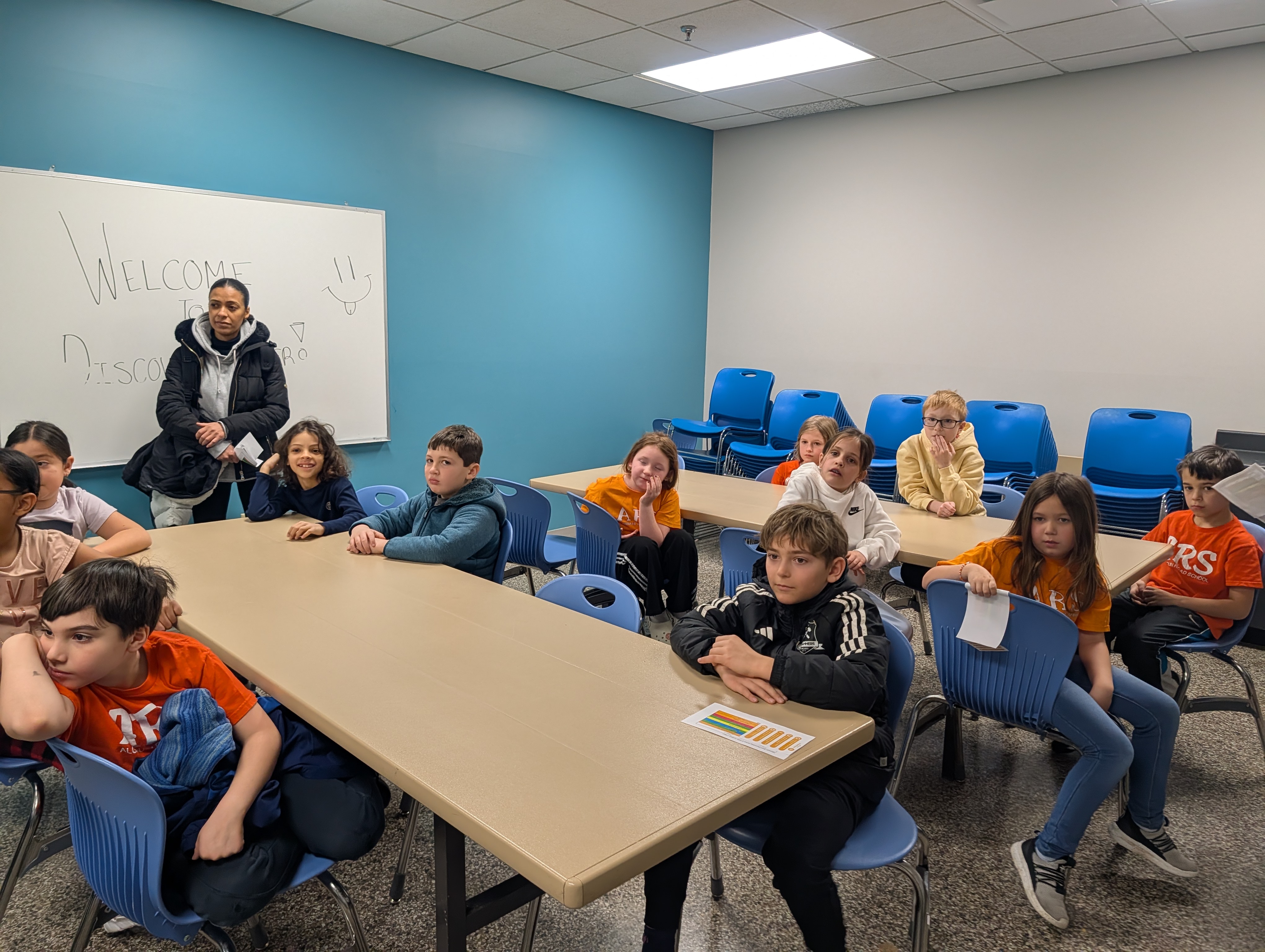 A group of students and an instructor are gathered in a classroom with tables and chairs.