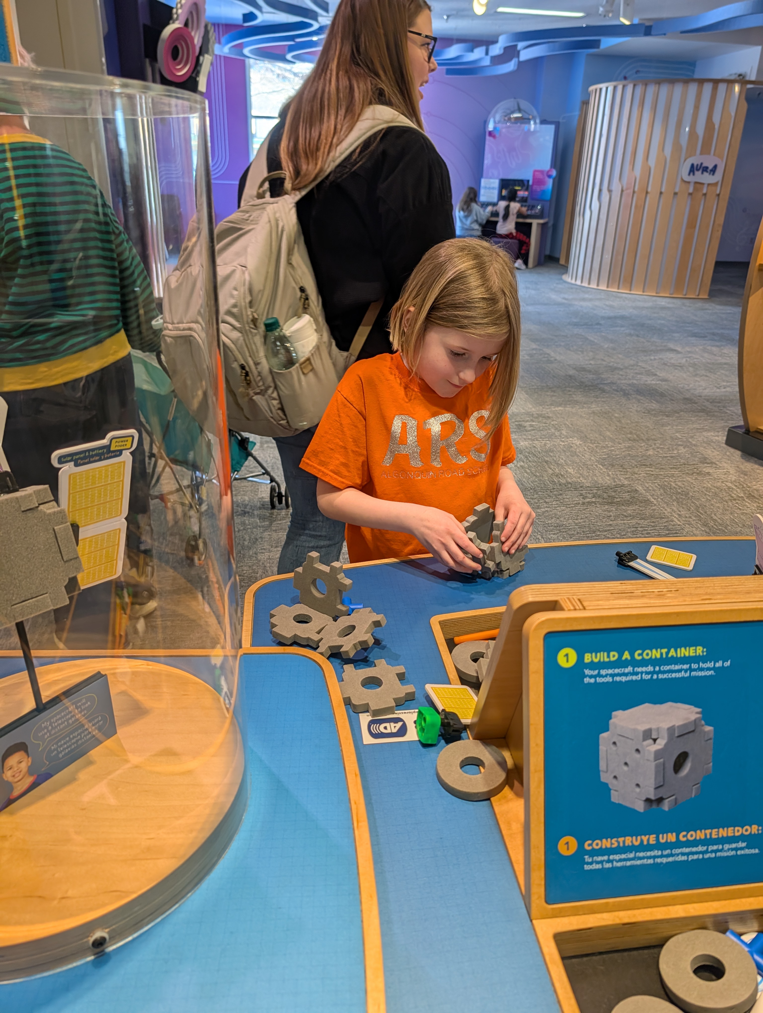 A young girl in an orange shirt concentrates on building with blocks at an interactive exhibit.