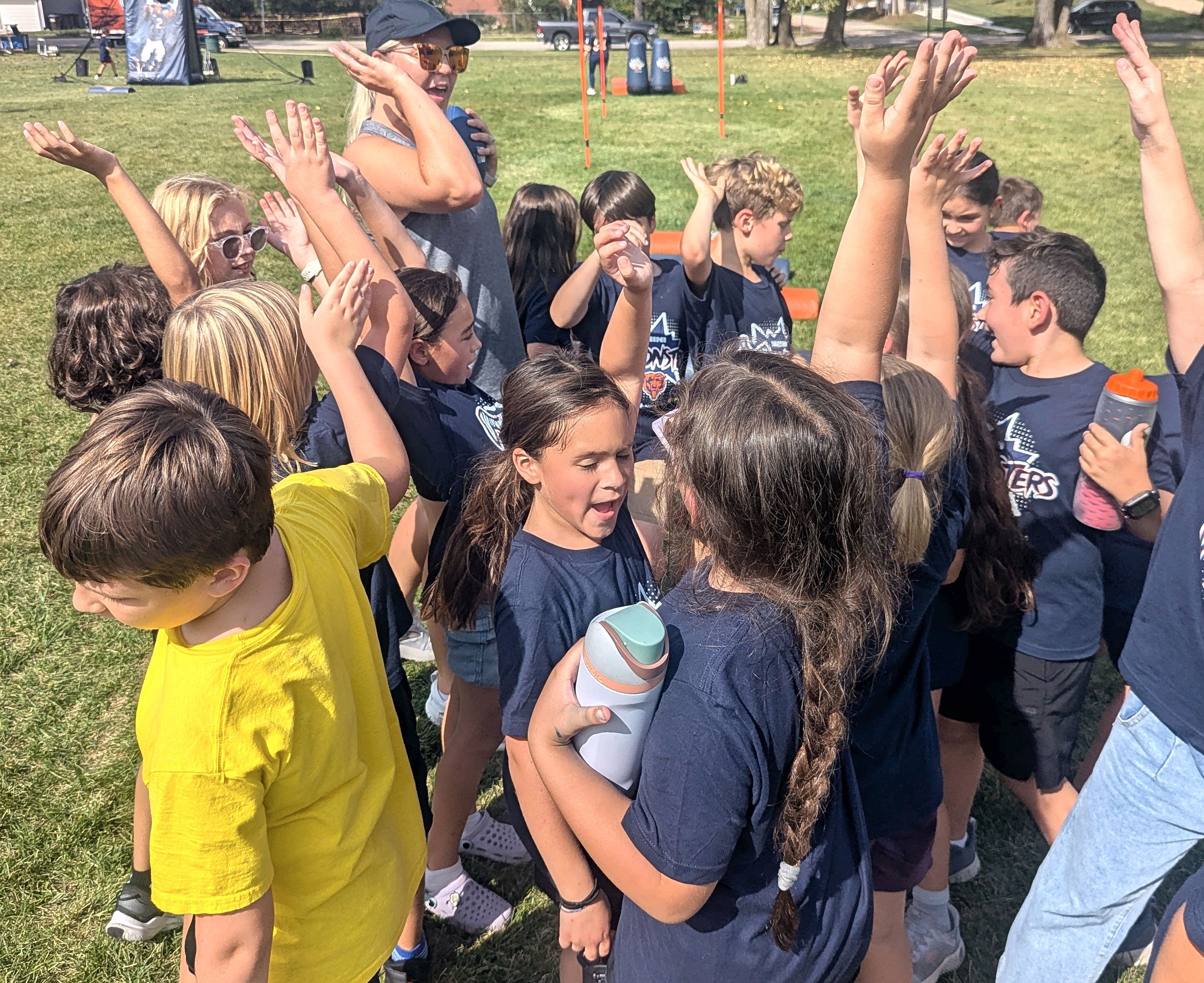 A group of children in navy shirts raise their hands and cheer outdoors.