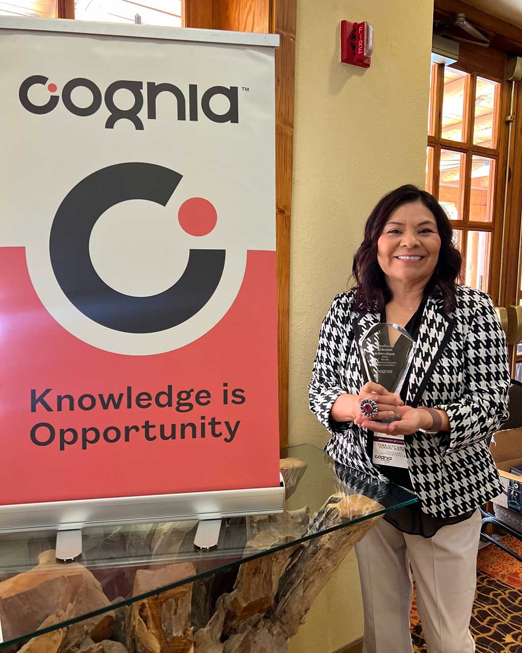 Dr. Bilagody smiles while standing next to a Cognia banner that reads "Knowledge is Opportunity" holding her award.