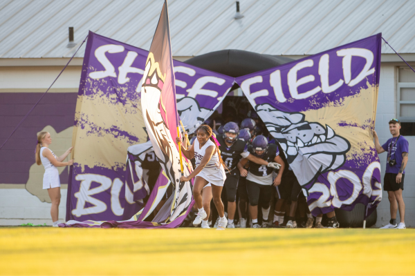 A football team runs through a purple and white banner that reads "SHEFFIELD BULLDOGS".