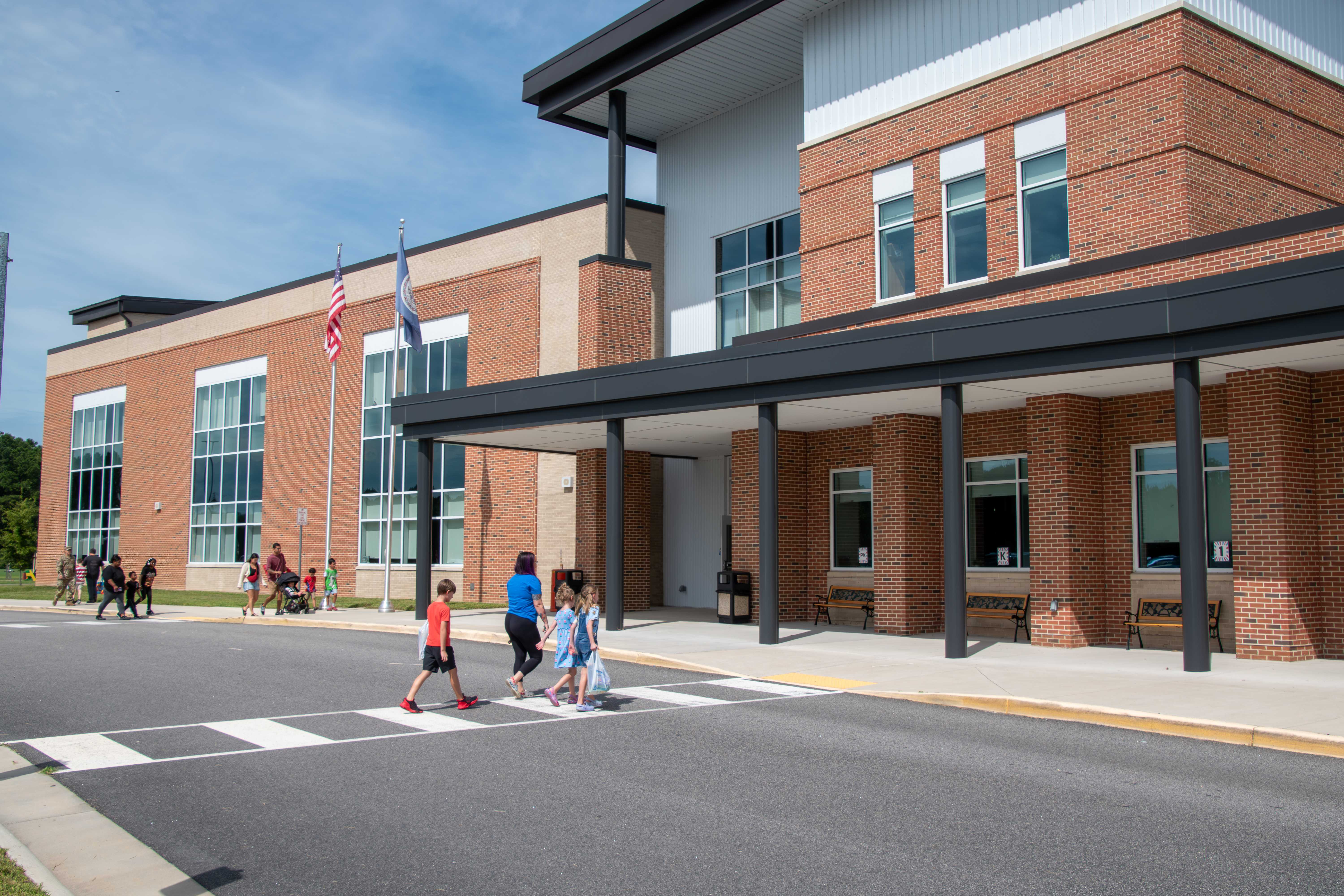 Children cross a crosswalk in front of a modern brick school building on a sunny day.