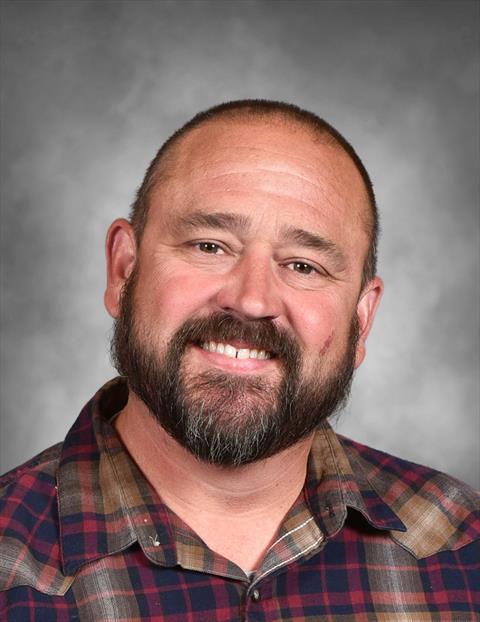 A portrait of a smiling man with a beard and a plaid shirt against a gray background.