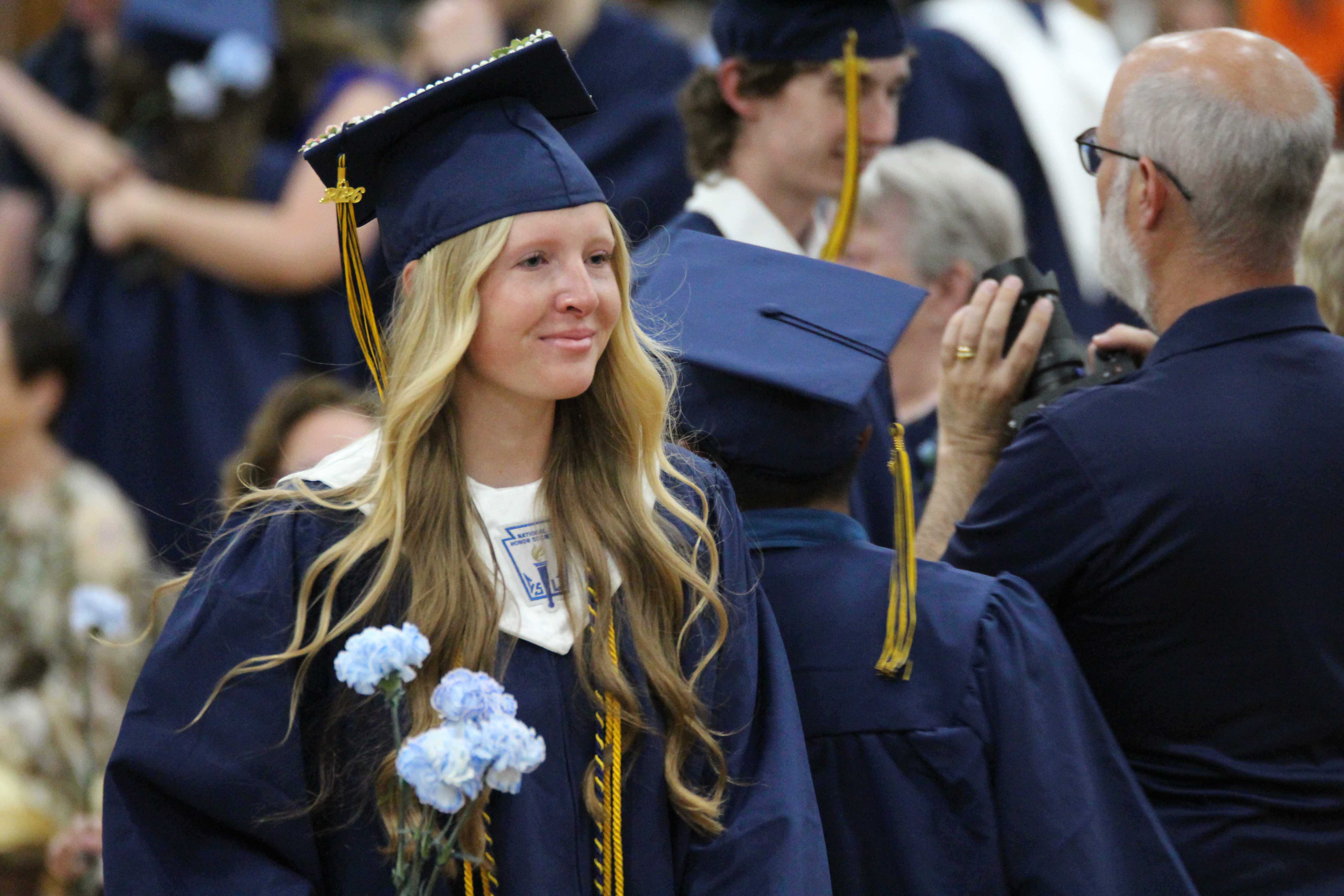 A young woman in a graduation cap and gown smiles, holding blue flowers.