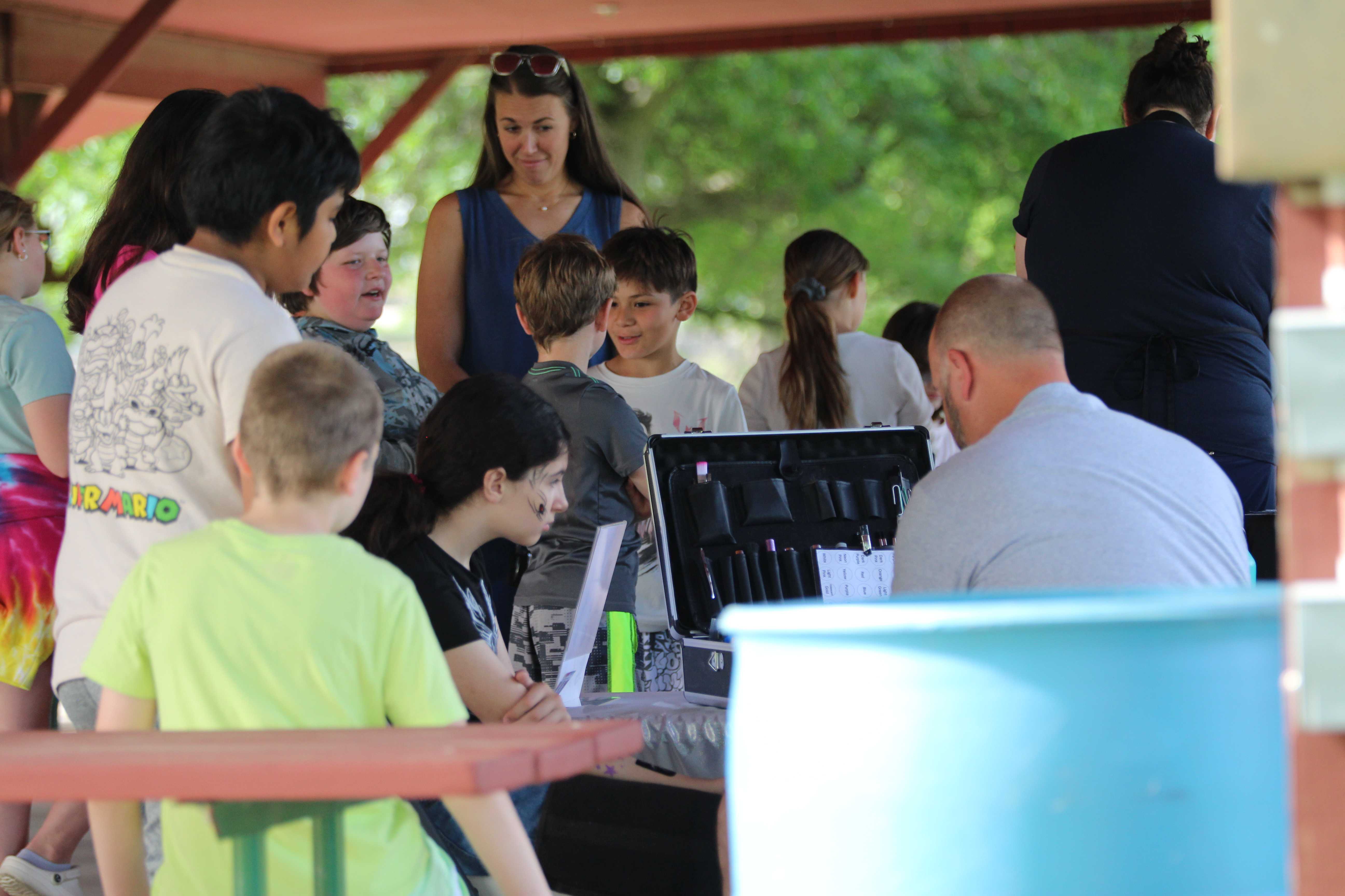 A group of children and adults gather under a covered pavilion, with some children looking at a case of items.