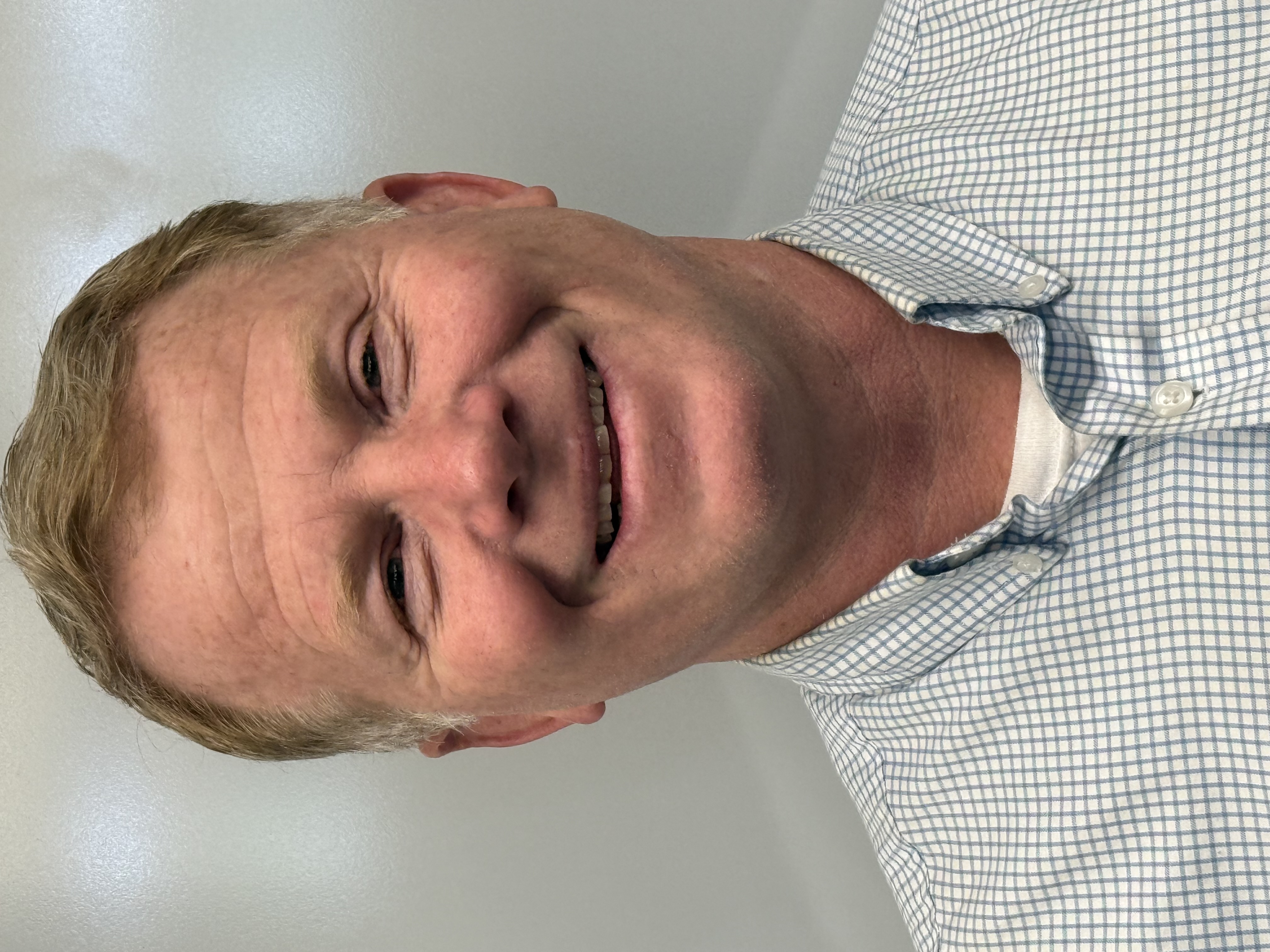 A smiling man with short blonde hair wears a light blue checkered shirt.