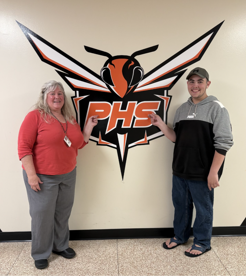 A woman and a young man stand in front of a large wall graphic featuring a stylized hornet and the letters 'PHS'.