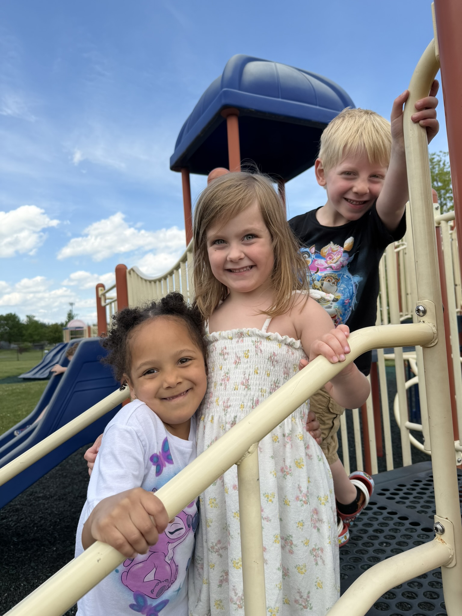 Three smiling children pose on a playground structure with a blue sky and clouds above.