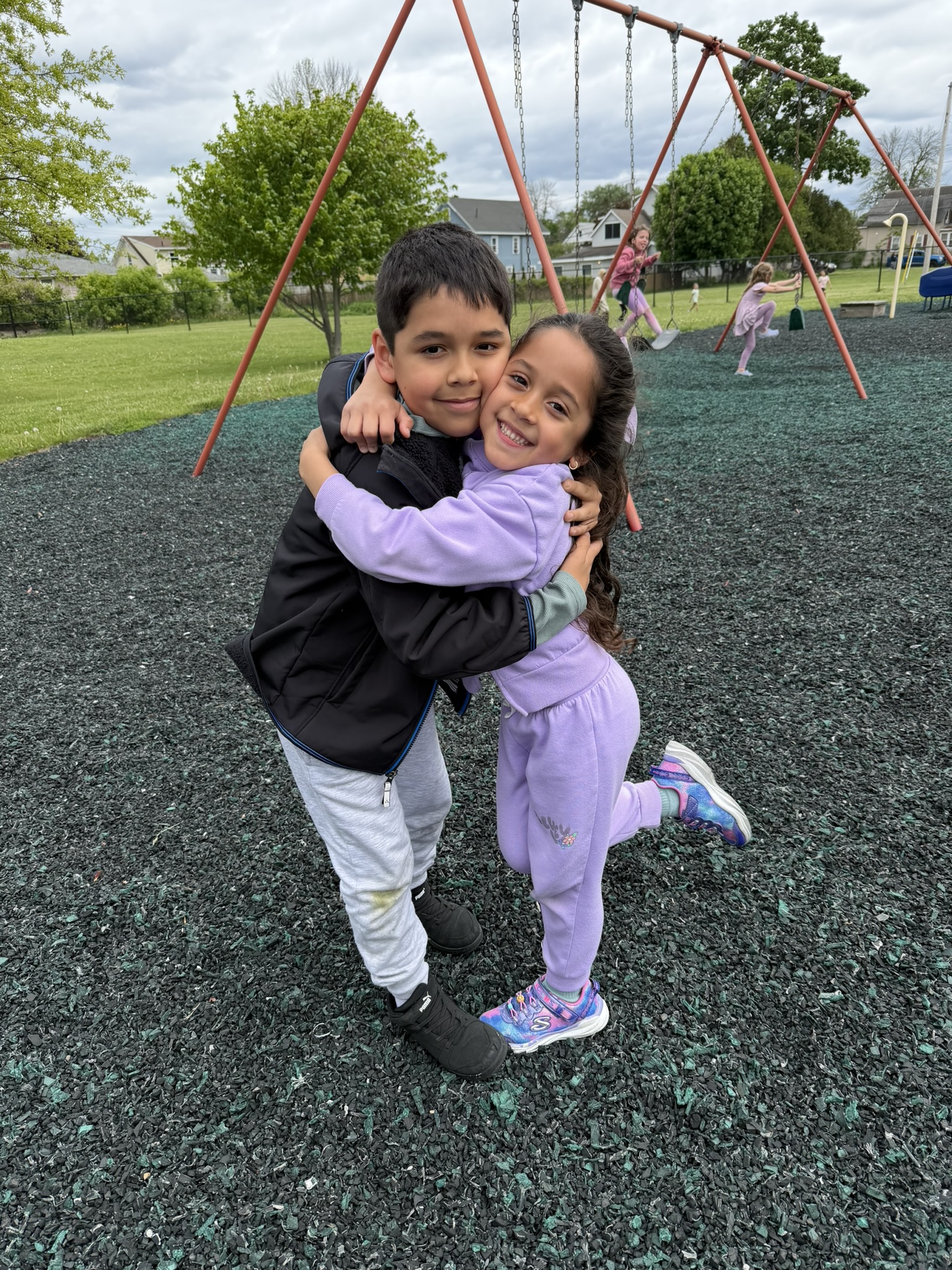 Two smiling children embrace warmly on a playground with swings in the background.