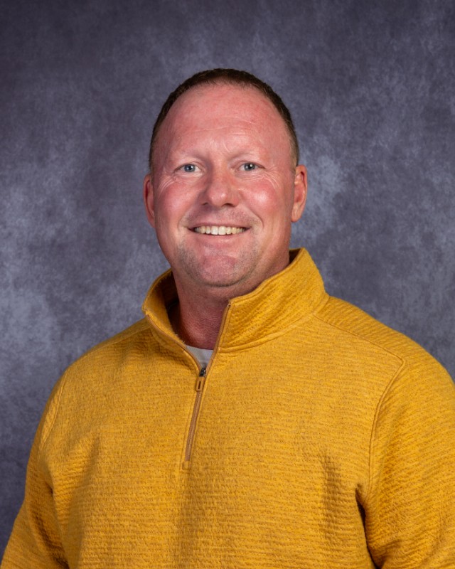 A smiling man wearing a yellow quarter-zip sweater poses in front of a mottled grey background.