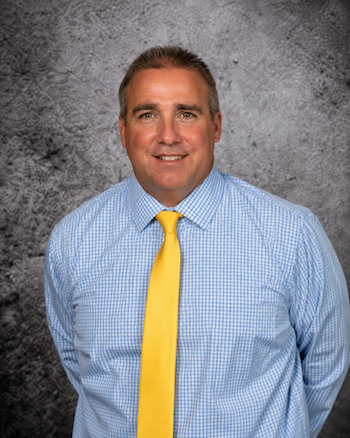 A man in a blue checkered shirt and yellow tie smiles at the camera.