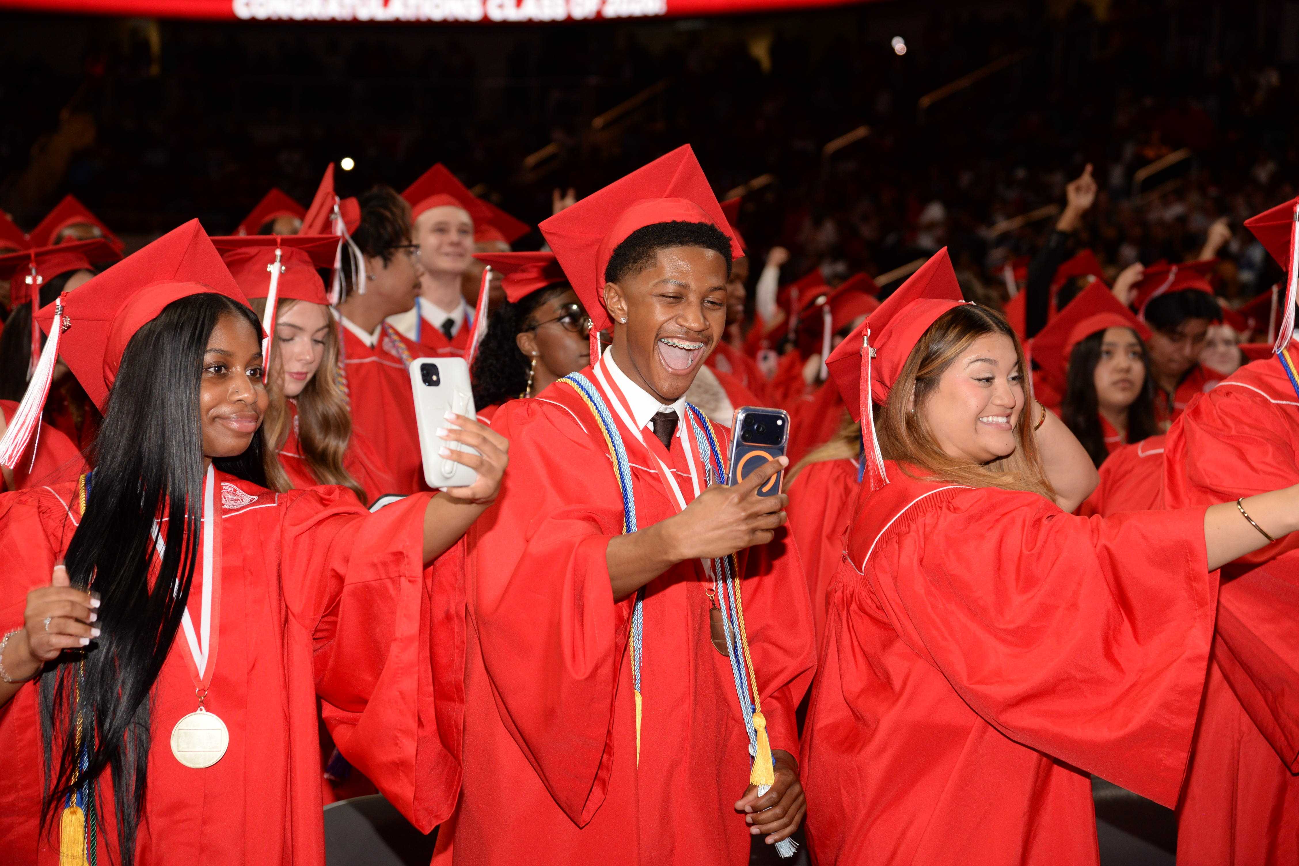 Graduates in red caps and gowns take selfies with their phones at a ceremony.