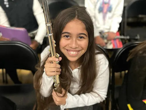 A young girl with long brown hair smiles brightly, holding a musical instrument.