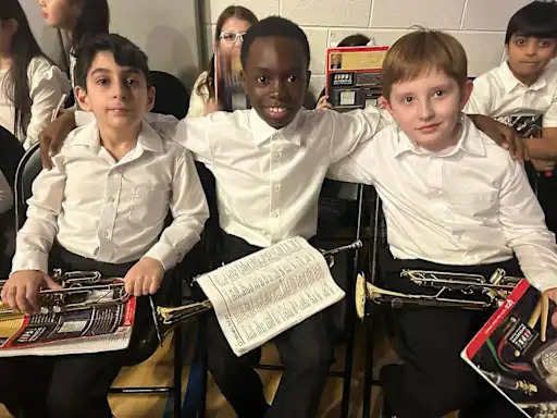 Three young boys in white shirts sit together, smiling and holding papers.
