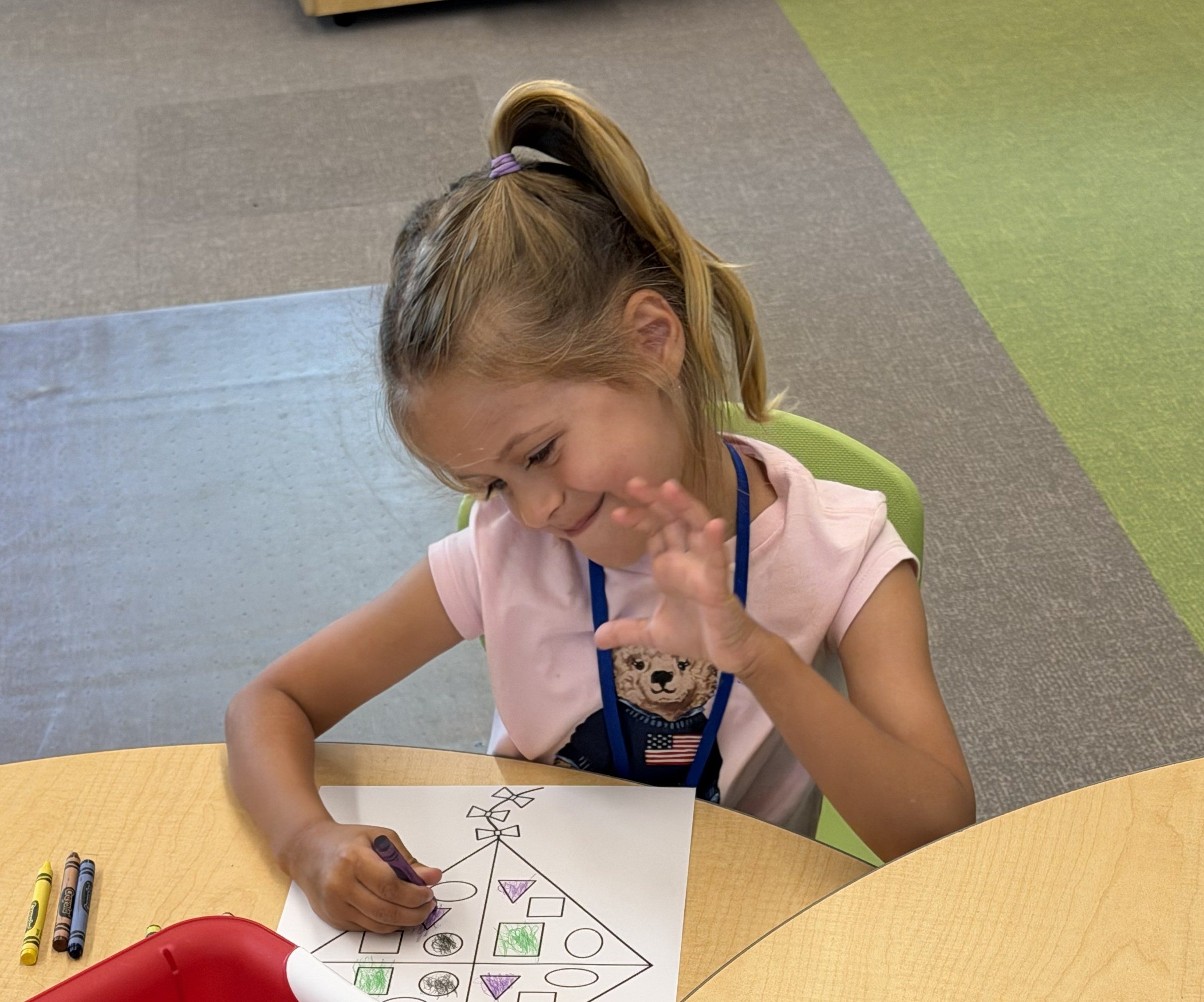 A young girl with blonde hair in a ponytail colors a picture at a table.