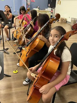 A young girl smiles while holding a cello bow during a music lesson.
