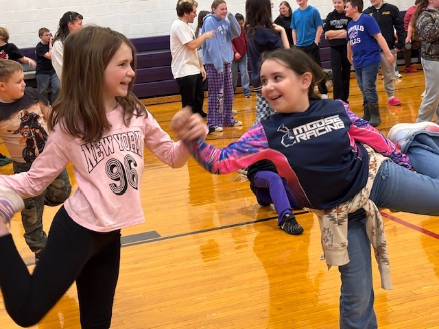 Two young girls hold hands and smile while dancing in a gymnasium.
