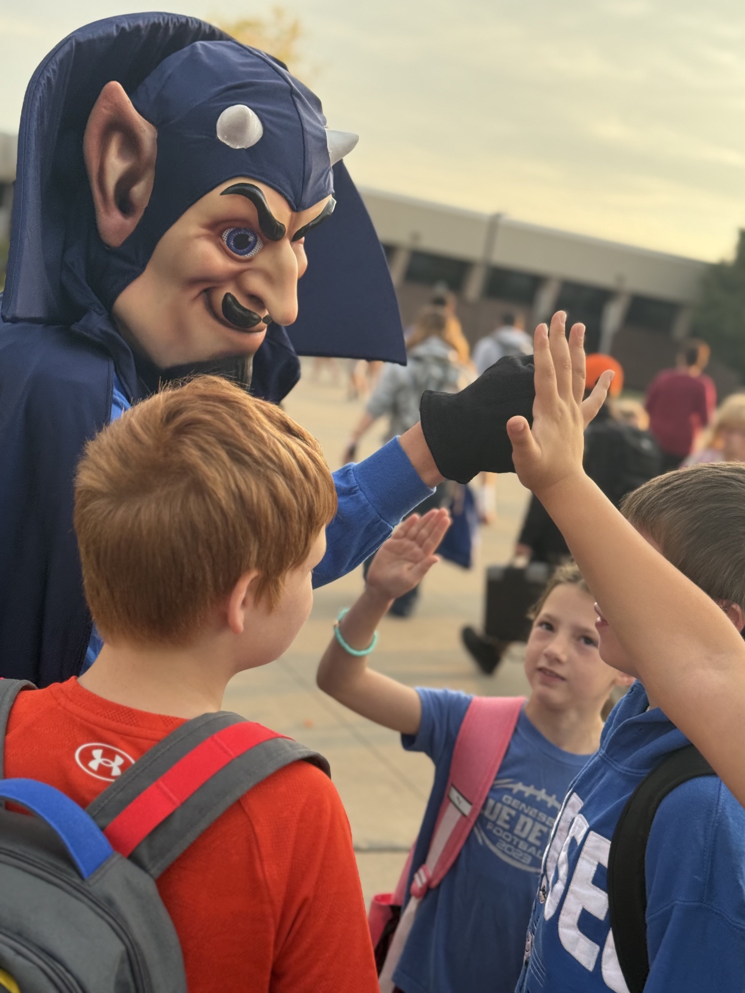 A mascot in a blue cape and mask gives a high-five to children.