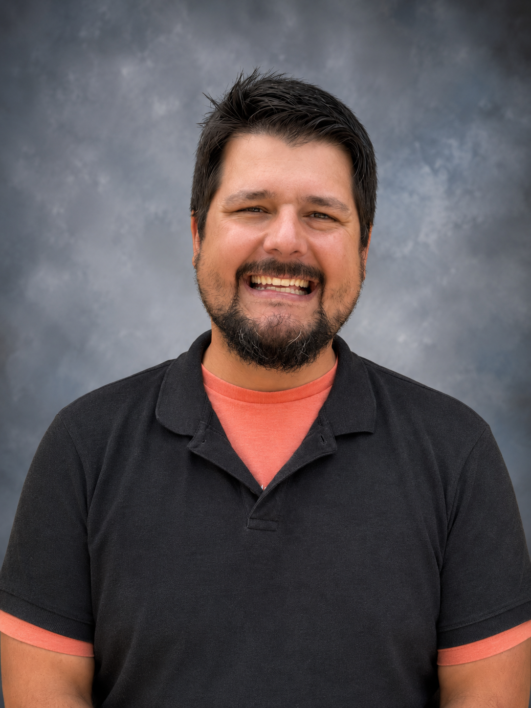 A man with dark hair and a beard smiles warmly at the camera, wearing a black polo shirt over an orange t-shirt.