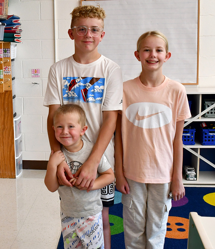 Three children stand together indoors, smiling while posing for a photo in a brightly lit room.