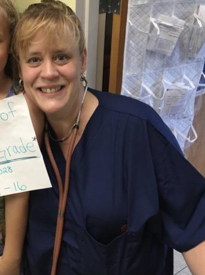 A smiling woman with short blonde hair wears dark blue scrubs and a stethoscope around her neck.