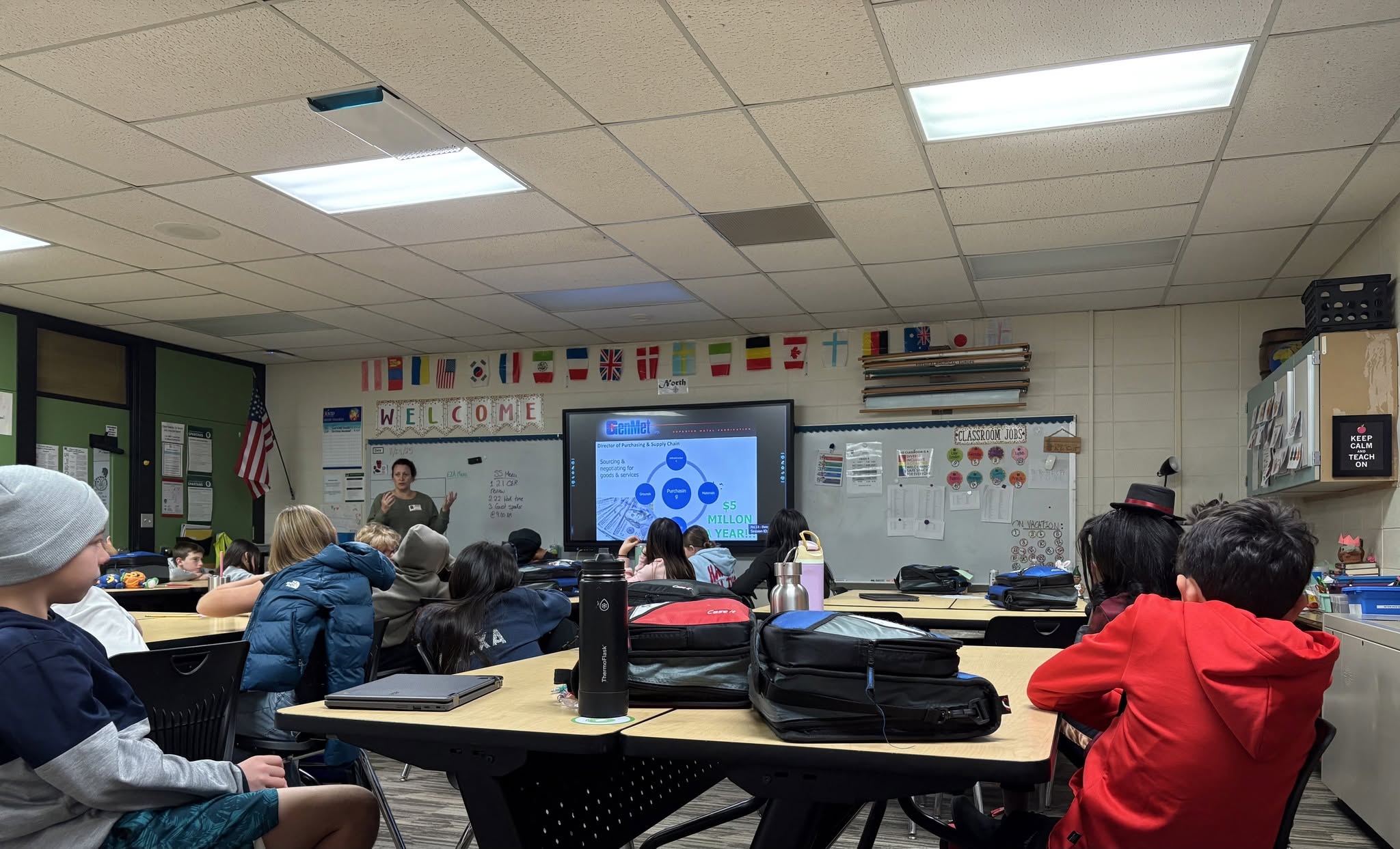 Students sit at desks in a classroom, facing a teacher presenting at a smartboard.