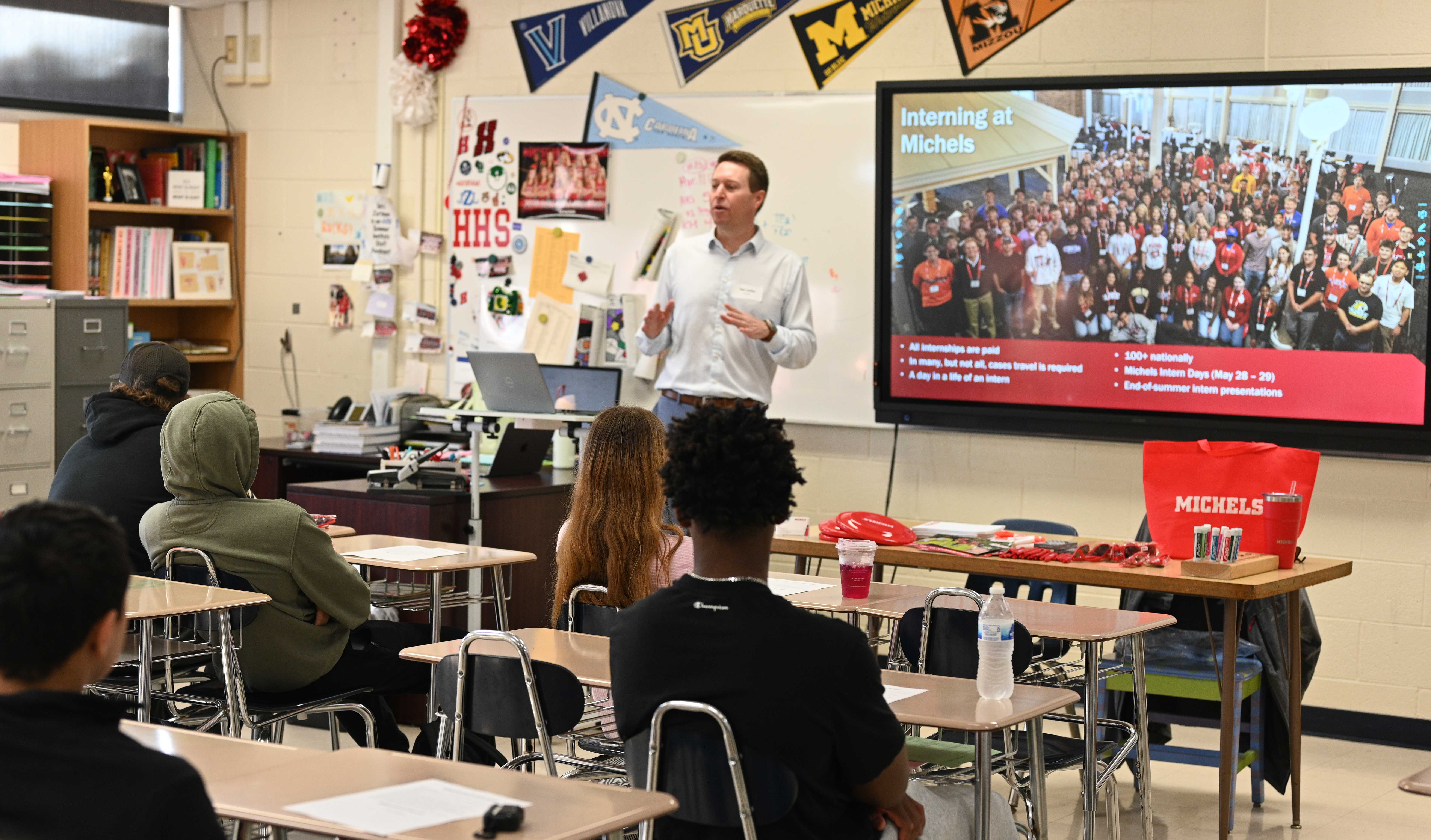 A teacher presents to a class of students in a classroom with a large screen displaying a group photo.